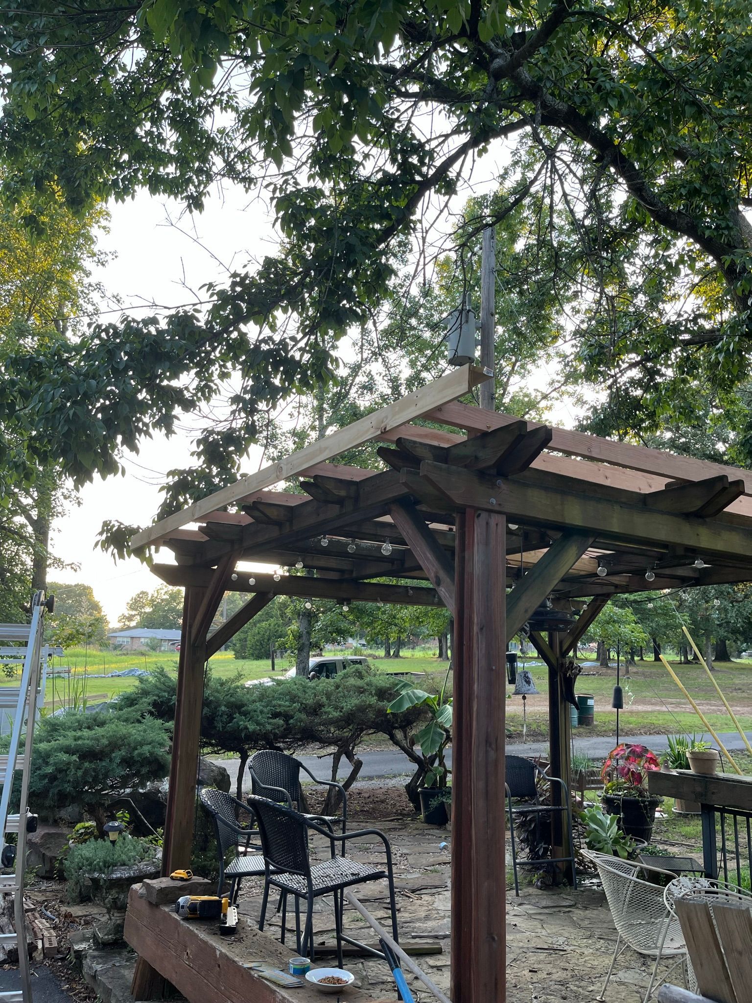 Pergola with antenna on top, chairs, plants. Green trees, sunny day.