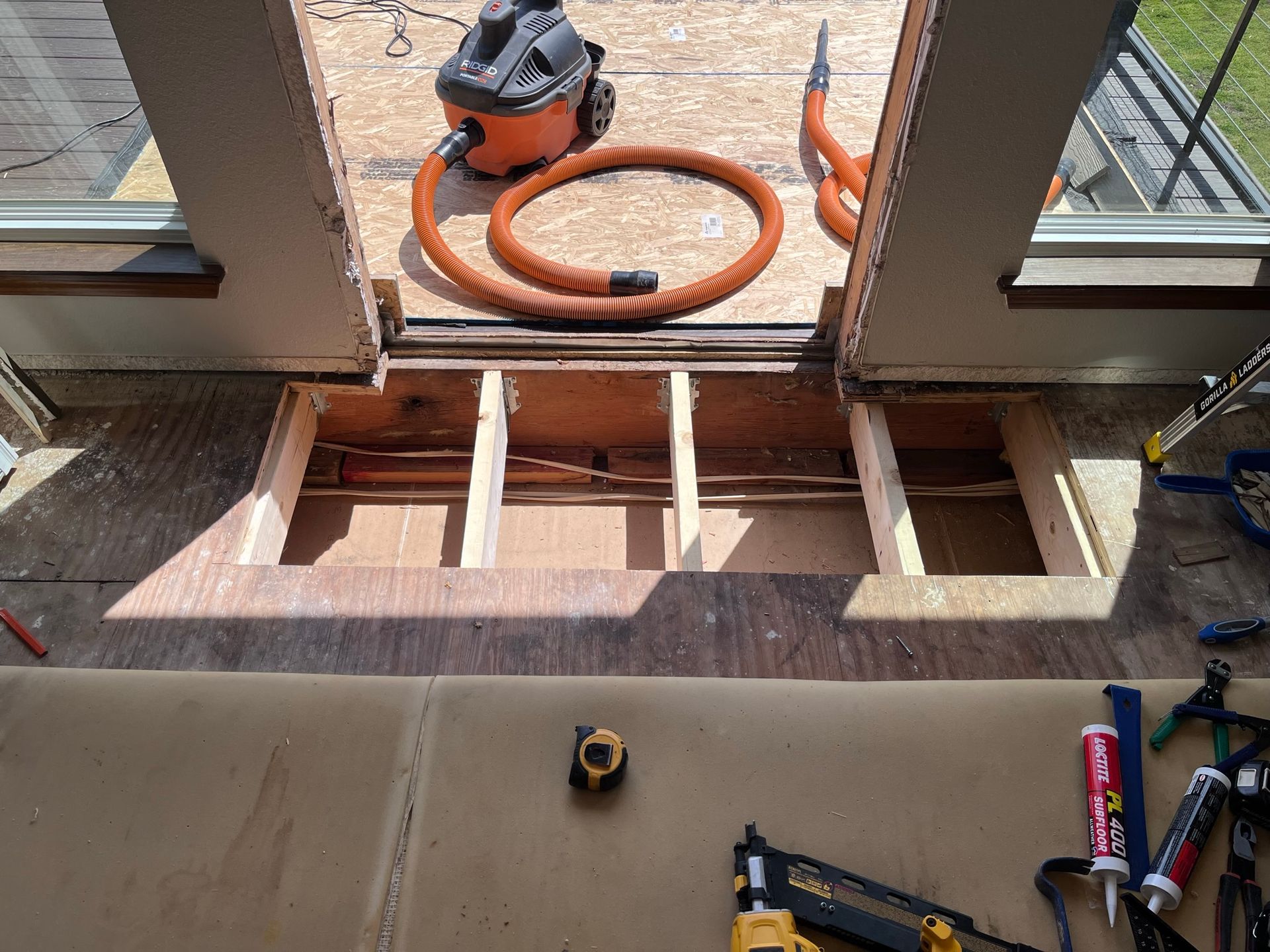 Interior view of exposed floor joists. Vacuum and tools on plywood, with sections removed.