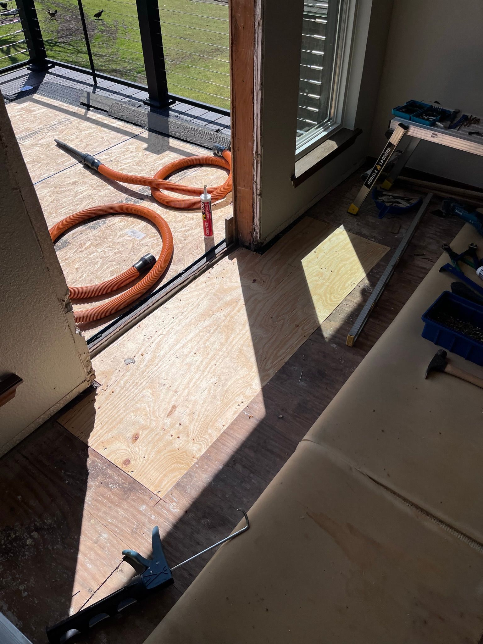 Plywood flooring being installed near an open doorway. An orange hose and tools are present. Sunlight streams in.