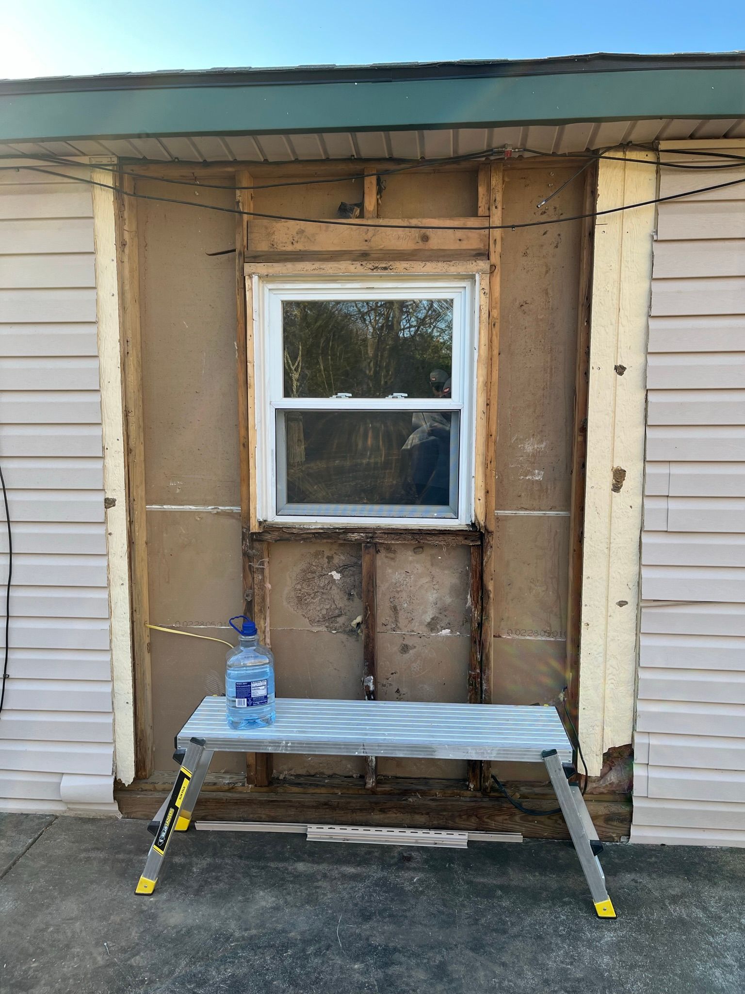Wall with exposed wood around a window, showing missing siding; a folding work table with a water bottle.