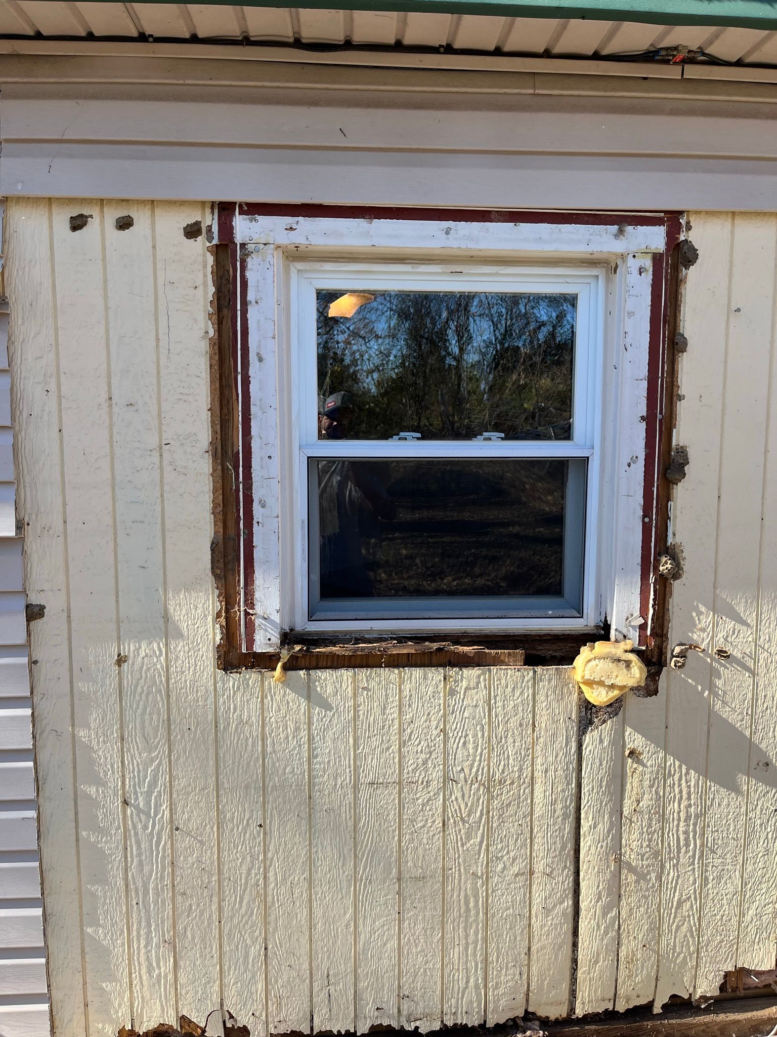 A window with a white frame in a weathered, yellow-sided building. Brown trim surrounds the window.