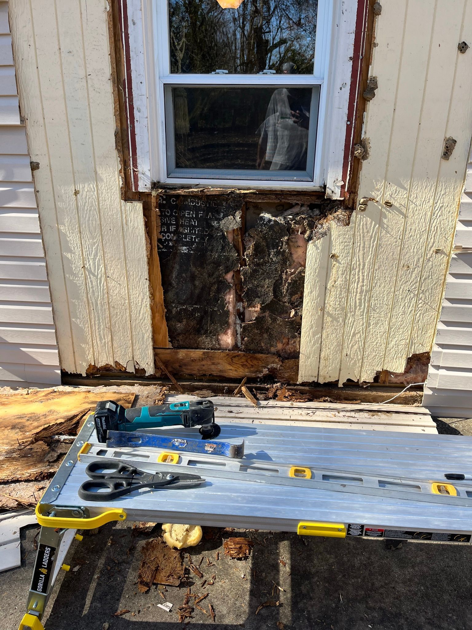 Window frame with damaged siding. Tools and supplies on a work table below.