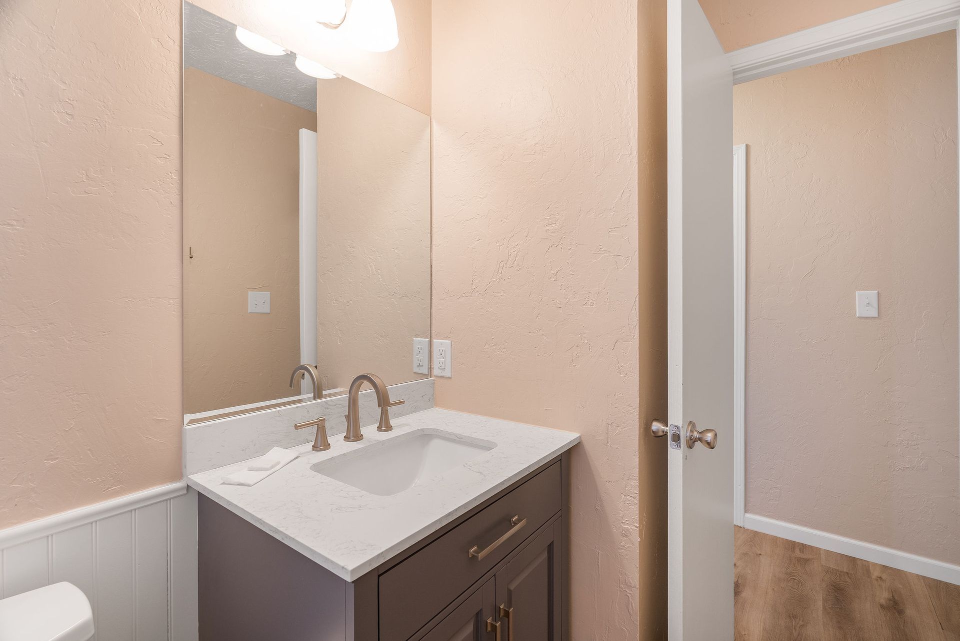 Bathroom with vanity, sink, mirror, and open door to toilet area. Peach-colored walls, wood-look floor.