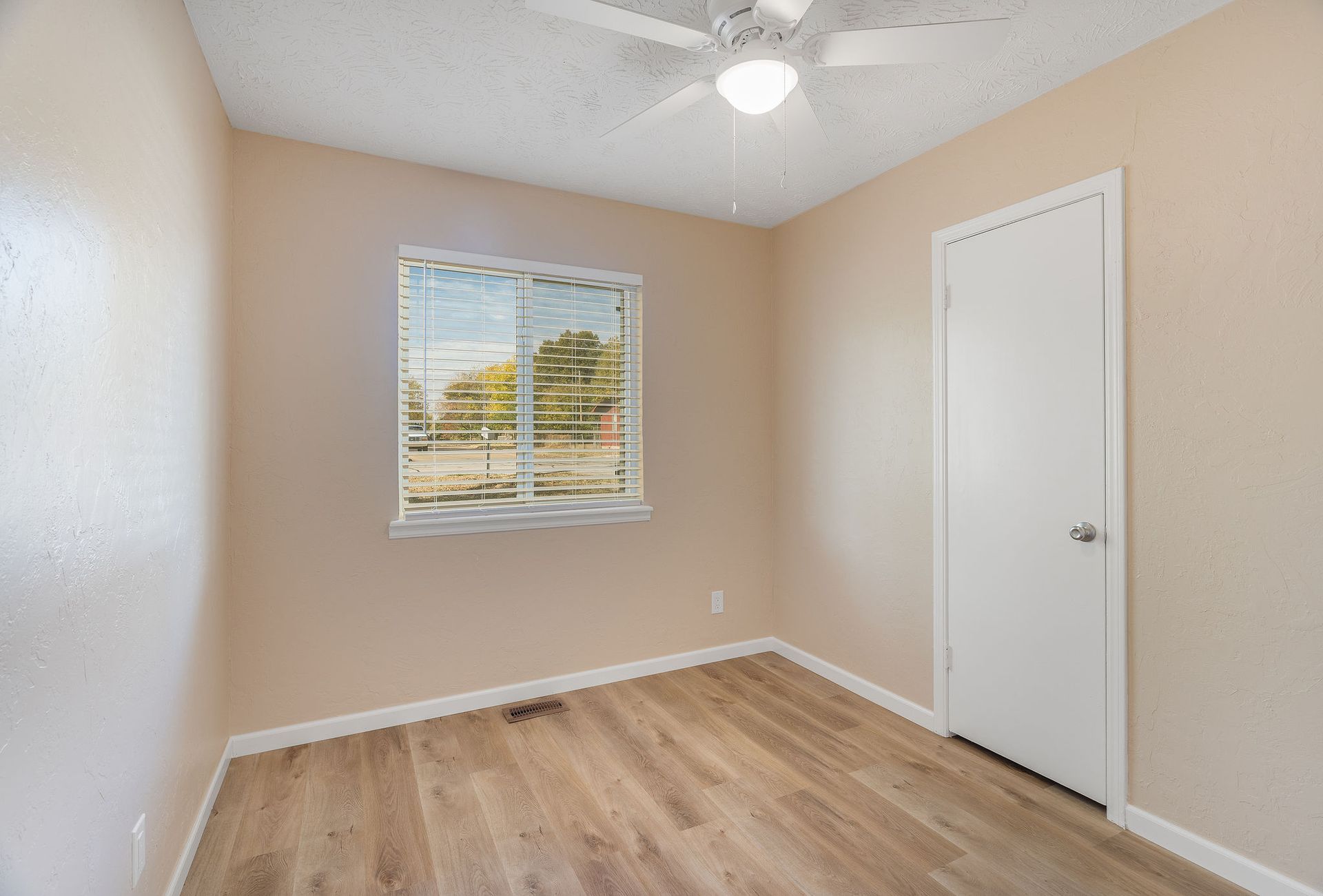 Empty room with beige walls, wood-look flooring, window, white door, and ceiling fan.