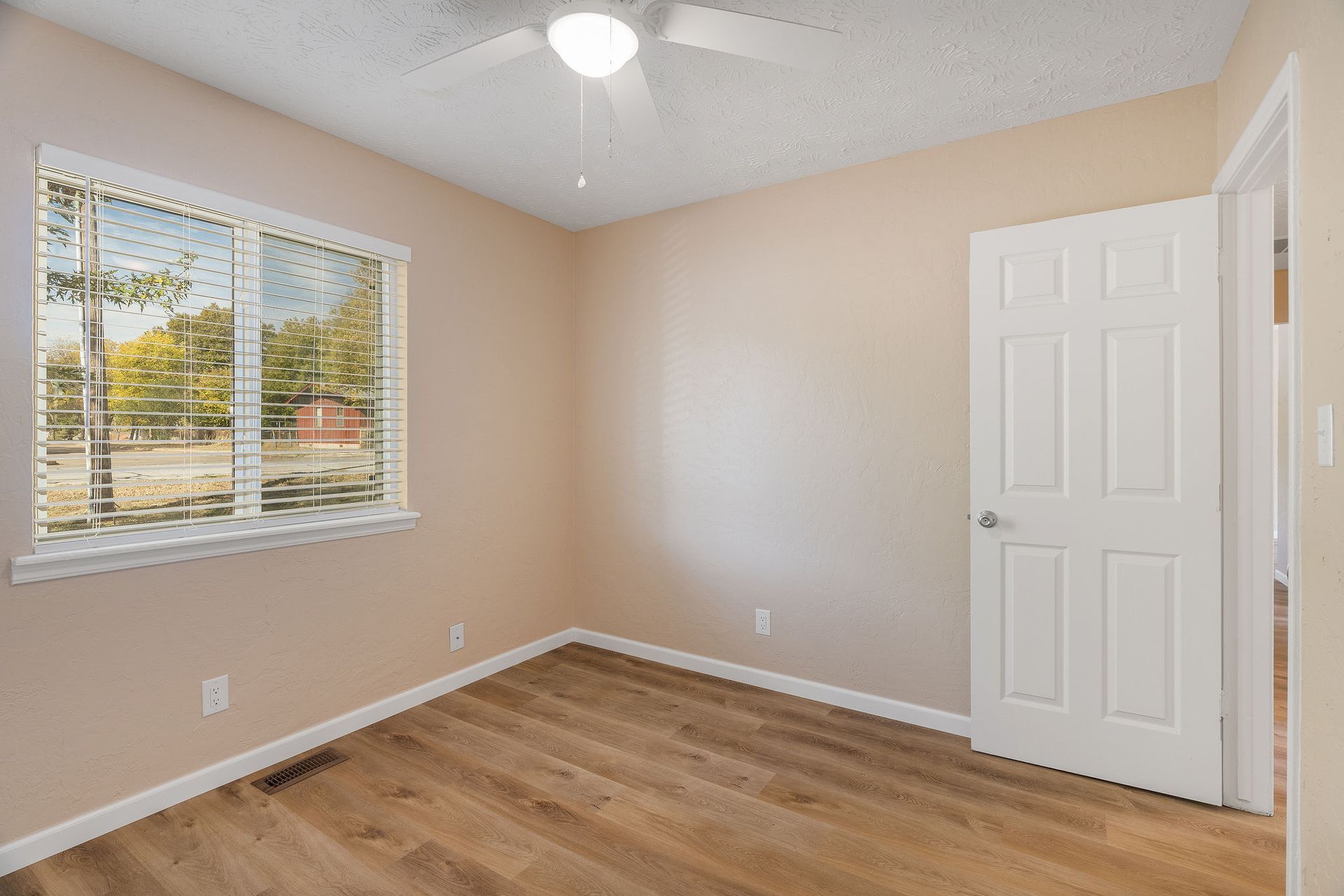 Empty bedroom with wood-look floor, window with blinds, beige walls, white door, and ceiling fan.