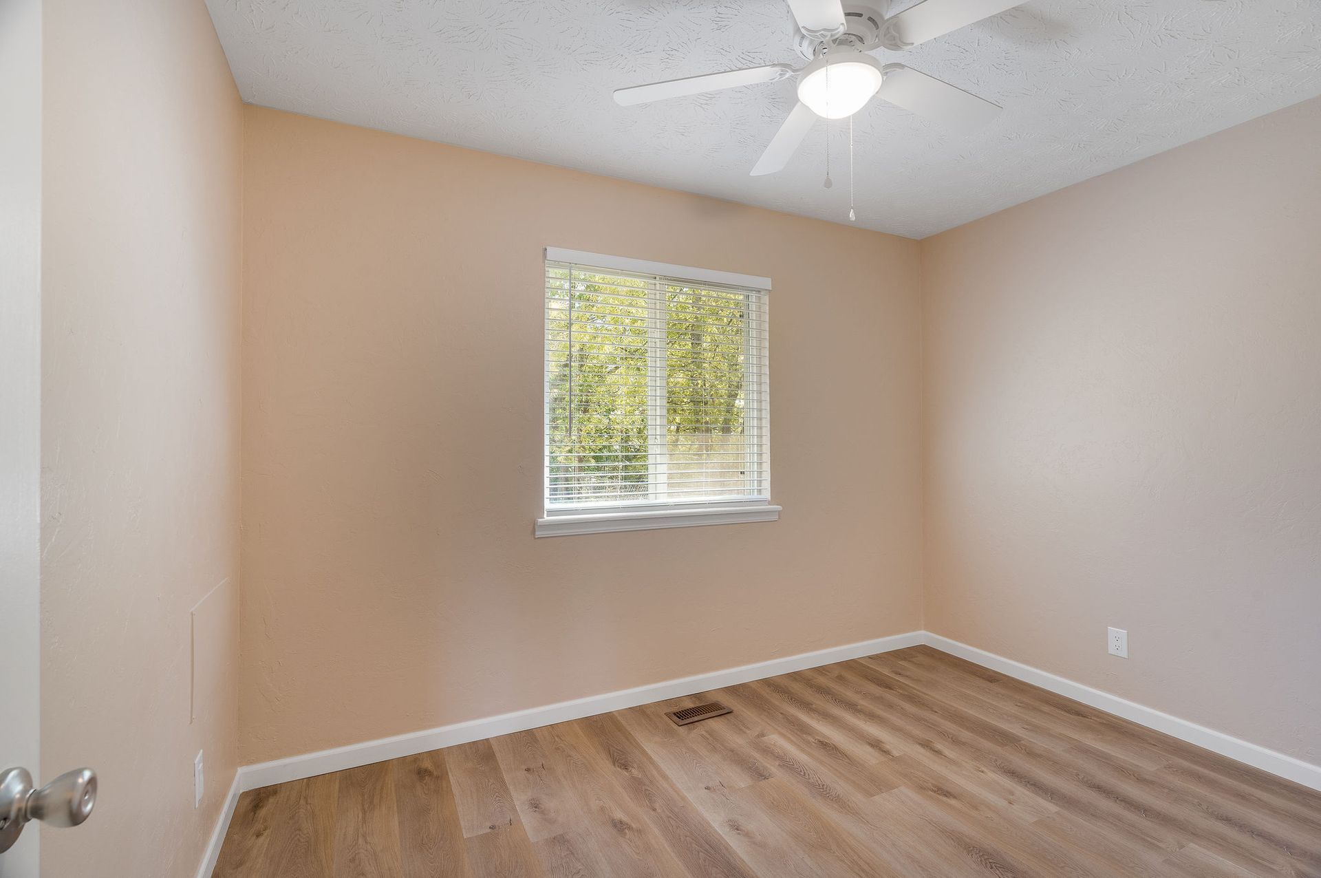 Empty beige bedroom with a window, ceiling fan, and wood-look flooring.