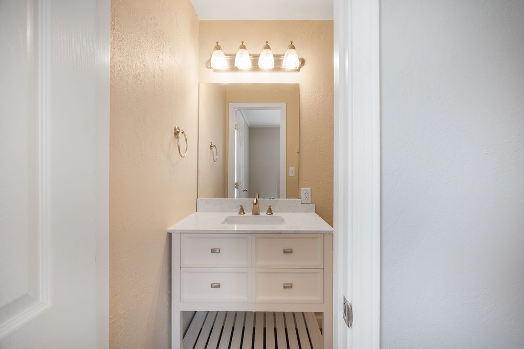 Small bathroom with white vanity, mirror, and gold fixtures. Light tan walls and black and white striped floor.