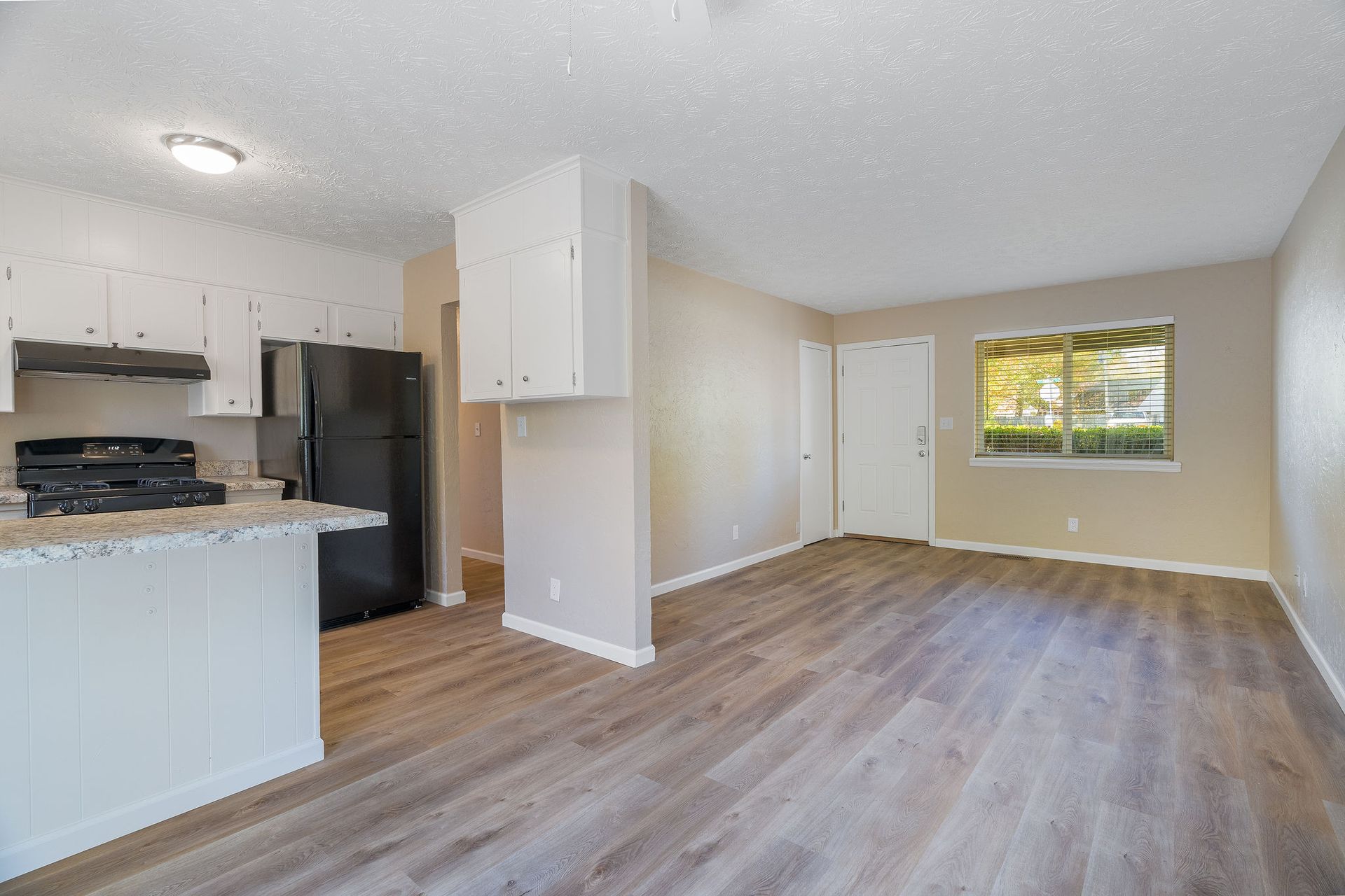 Kitchen and living area of an apartment with white cabinets, gray countertops, black appliances, and gray wood flooring.