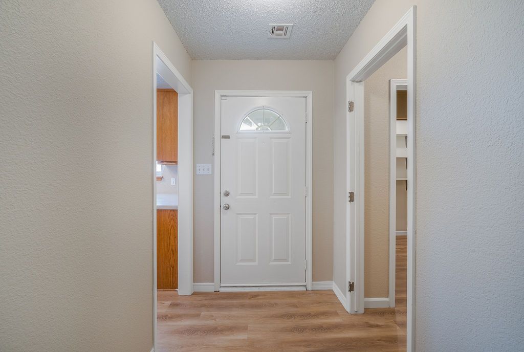 Hallway with white door and doorways to kitchen and room with shelves; light wood-look floor.