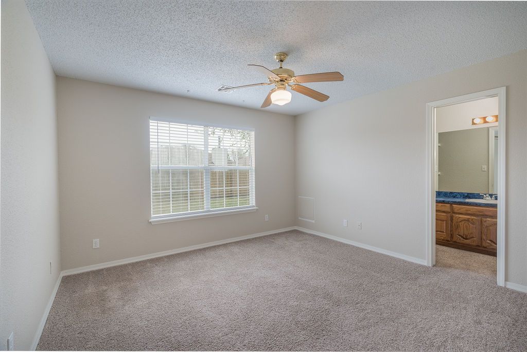 Empty bedroom with beige walls, carpet, window, and a ceiling fan. Bathroom visible in doorway.