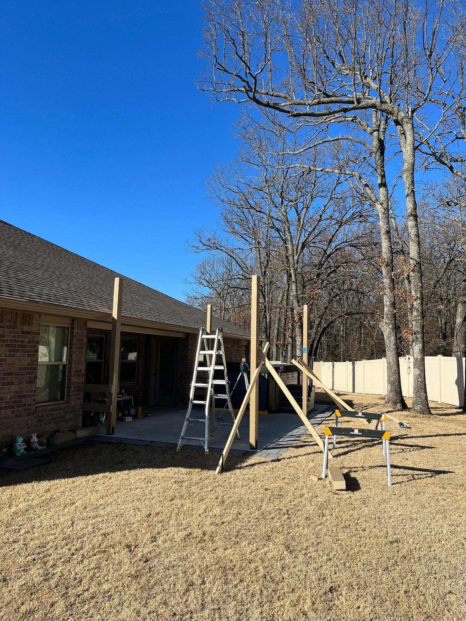 Construction of a patio cover: Wooden posts, ladders, and sawhorses in a backyard. Clear blue sky.