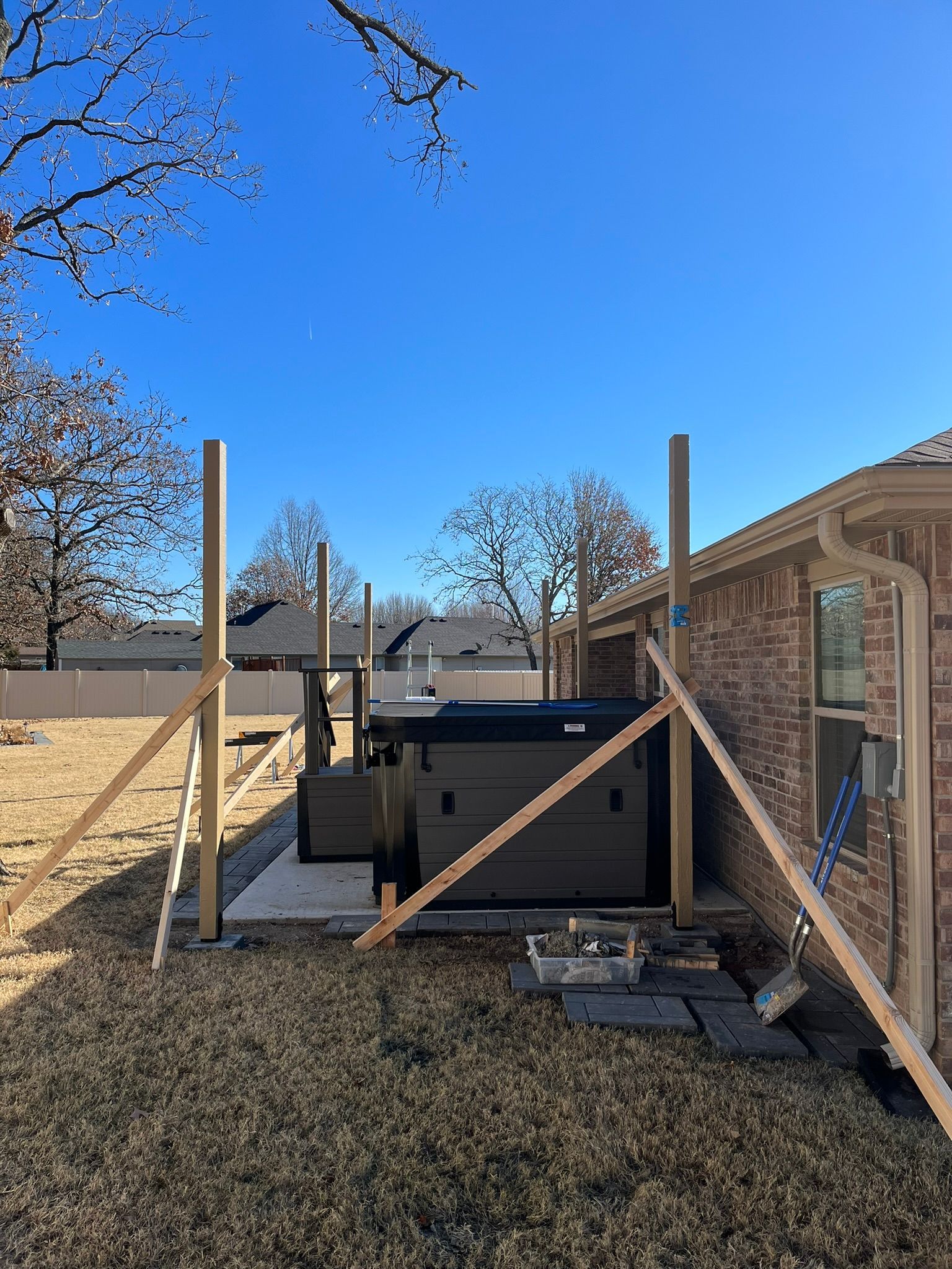 A hot tub installation in a backyard, supported by wooden posts and braces, against a brick home and clear blue sky.