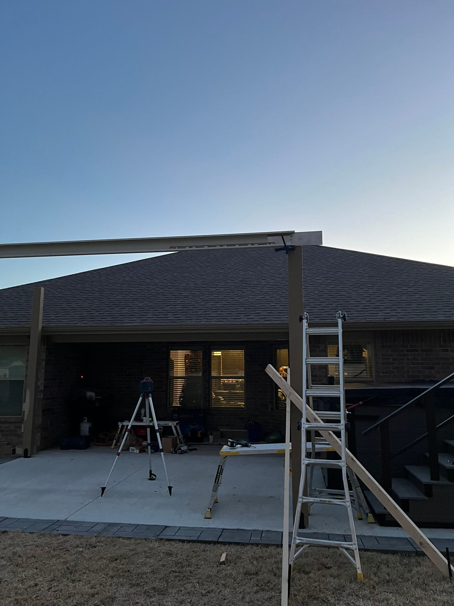 Backyard with construction: wooden beams, ladder, tripod, and a house with a tiled roof. Evening sky.