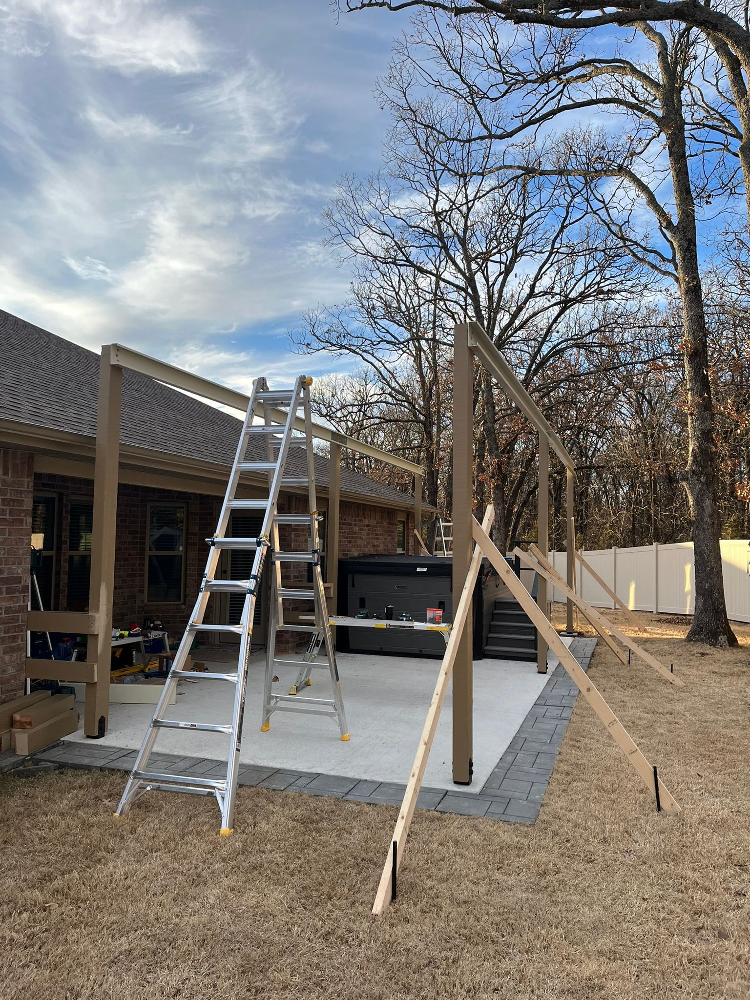 Construction of a patio cover: ladder, wooden beams, hot tub, and house in a yard on a cloudy day.