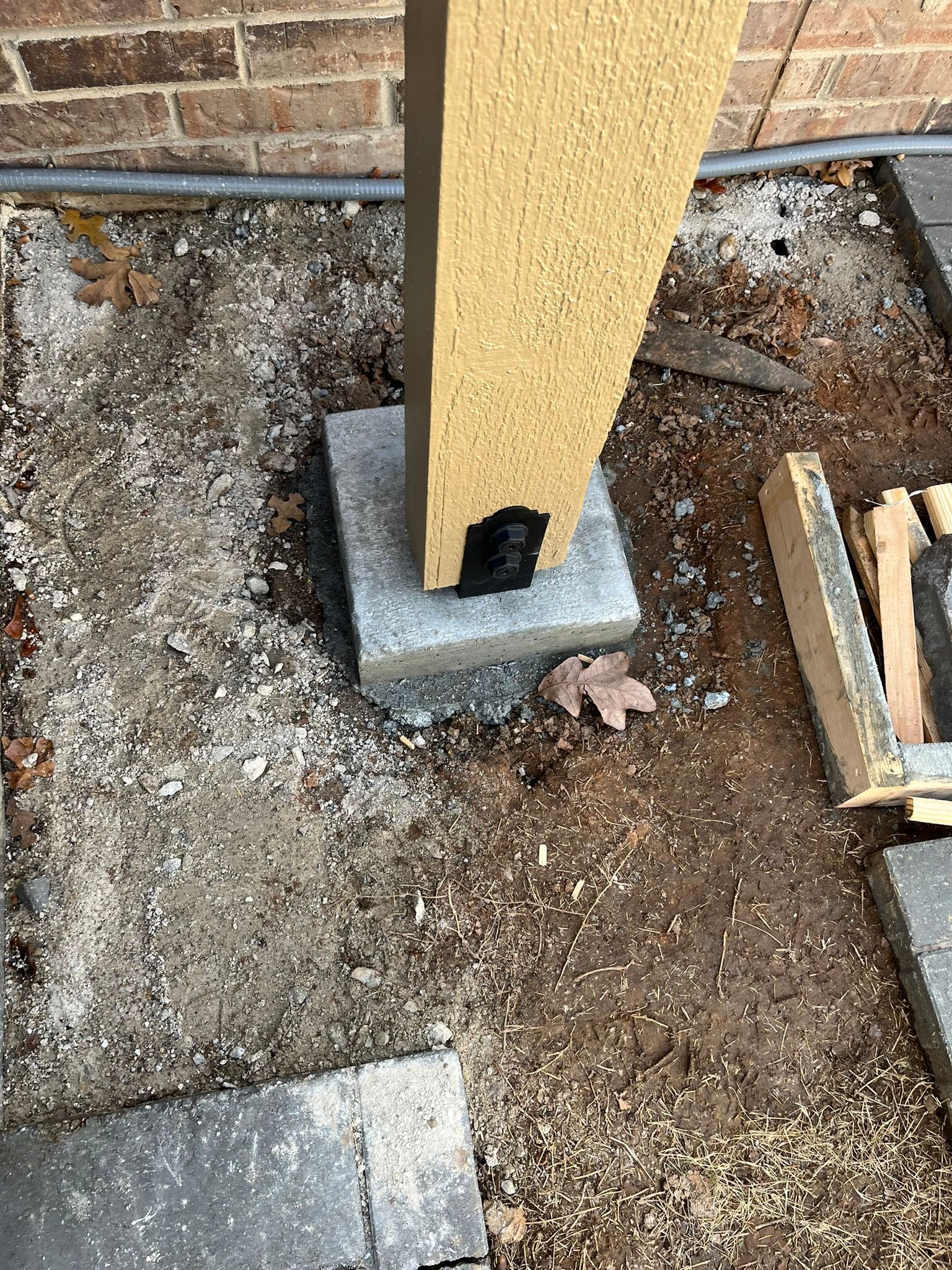 Concrete pillar base on gravel and soil with a nearby brick wall.
