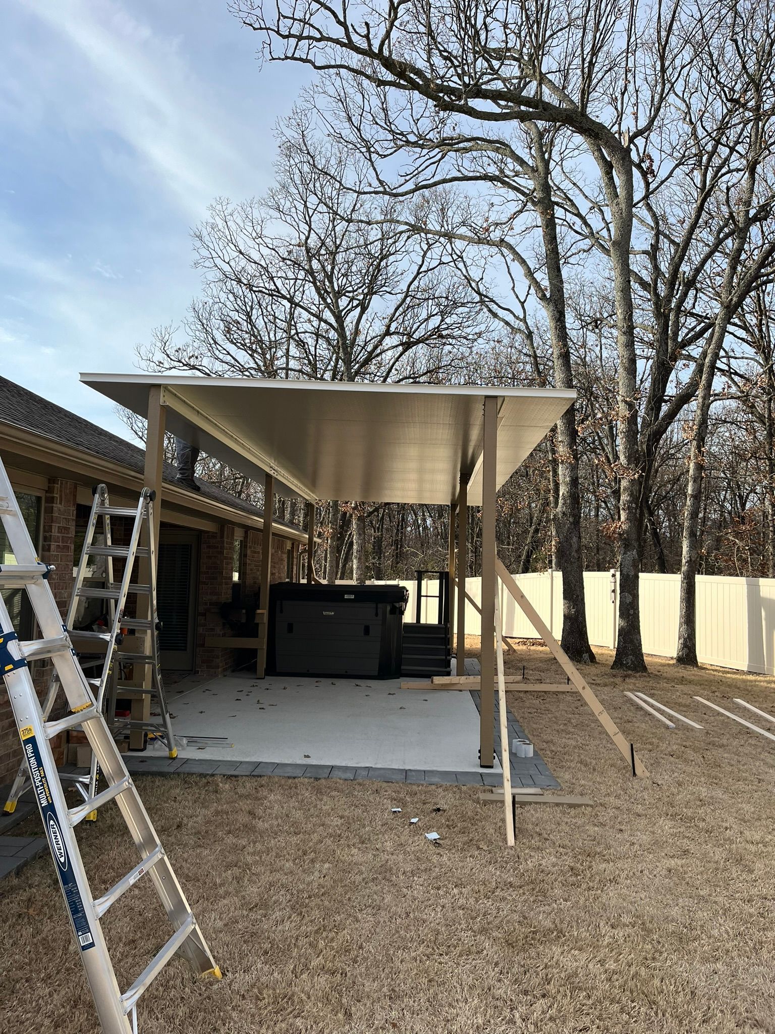 A partially constructed patio cover in a backyard with a hot tub and a ladder leaning against a brick house.