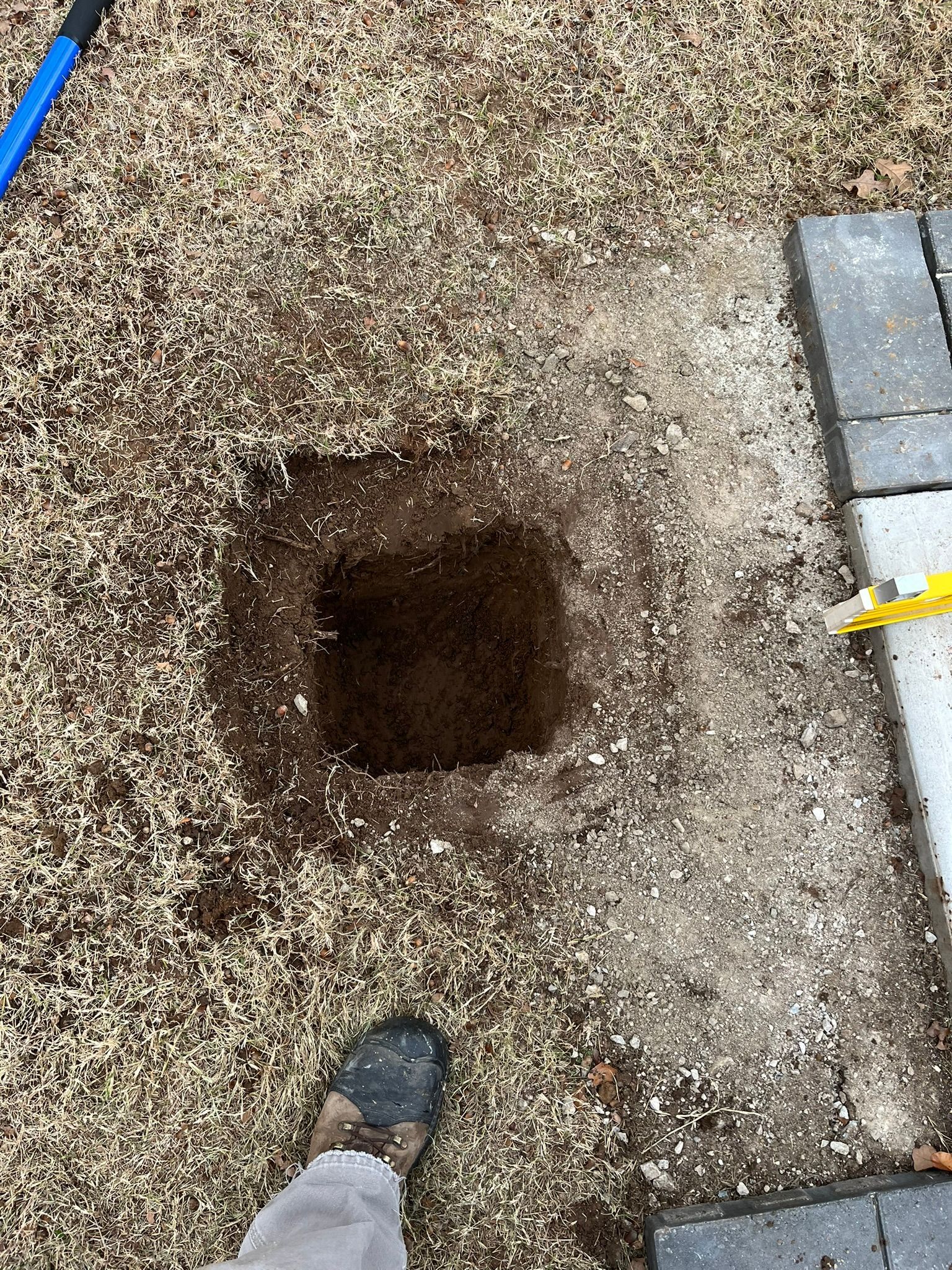 Square hole dug in a yard with gravel and a foot visible. Nearby are pavers and a shovel.