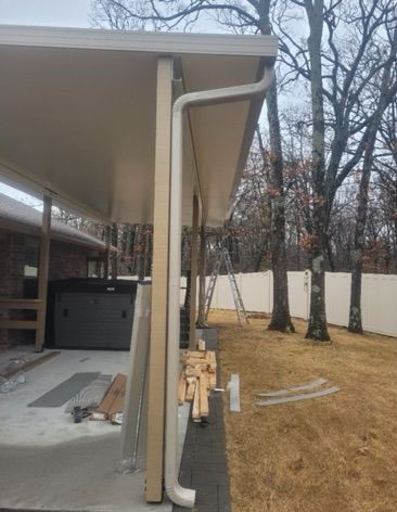 Beige-roofed carport with gutter, side view. Construction materials on ground. Trees and white fence in background.