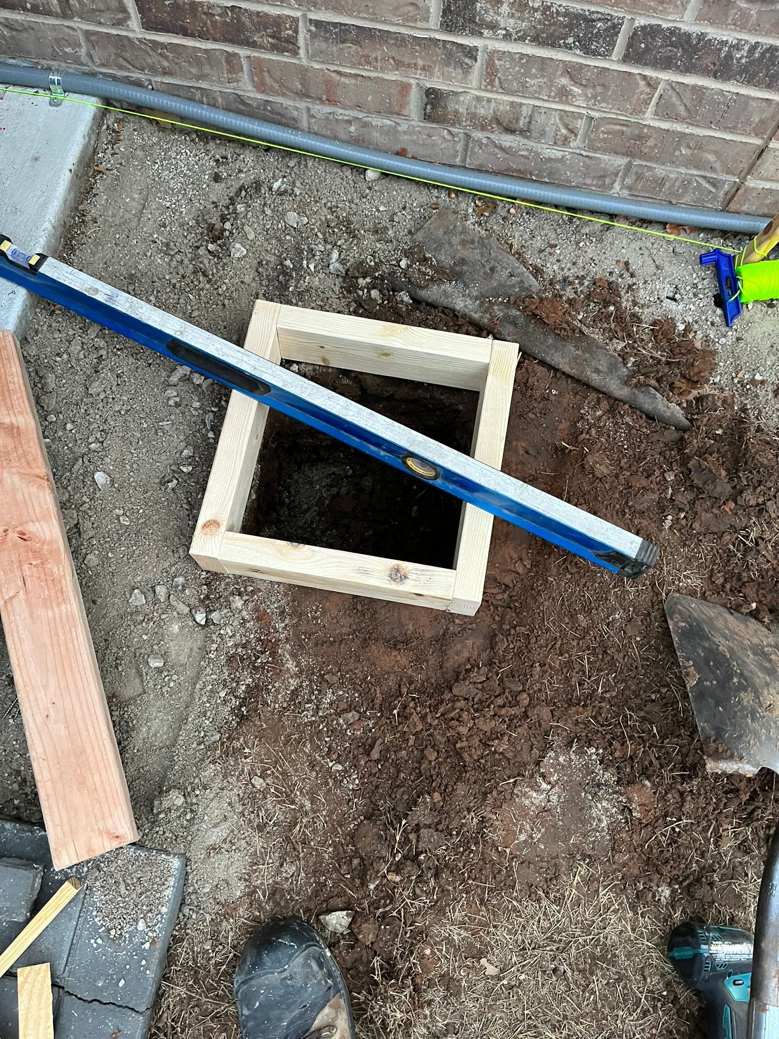 Wooden frame in a dirt hole, level resting on top, near brick wall and pathway.