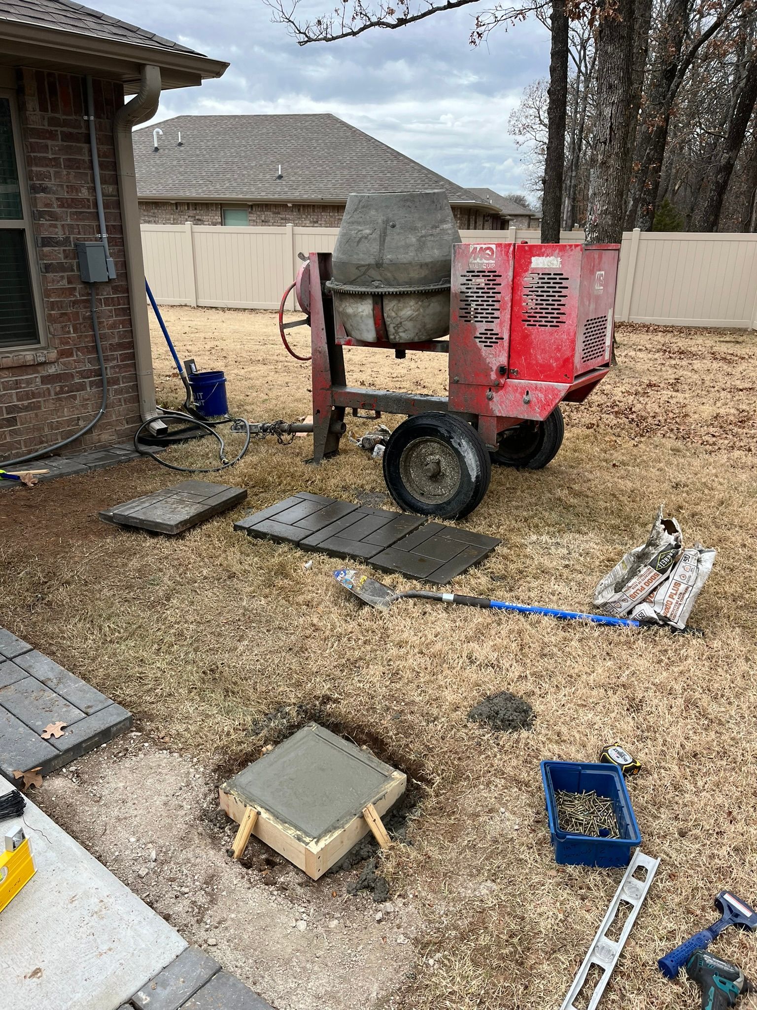 Concrete mixer and tools on grass, with partially poured concrete stepping stones near a brick house.