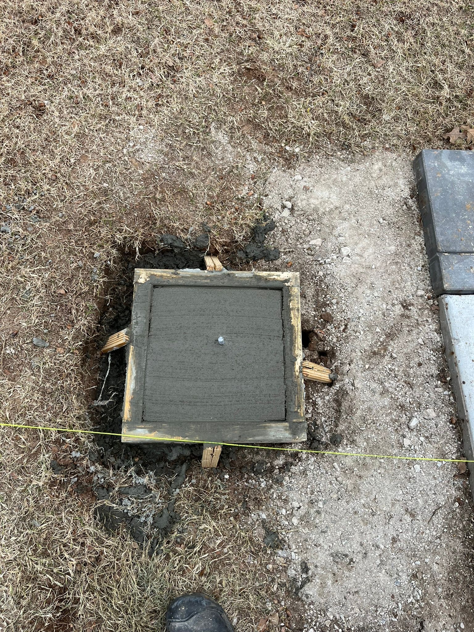A square wooden frame filled with wet concrete, set on gravel, next to paving stones.