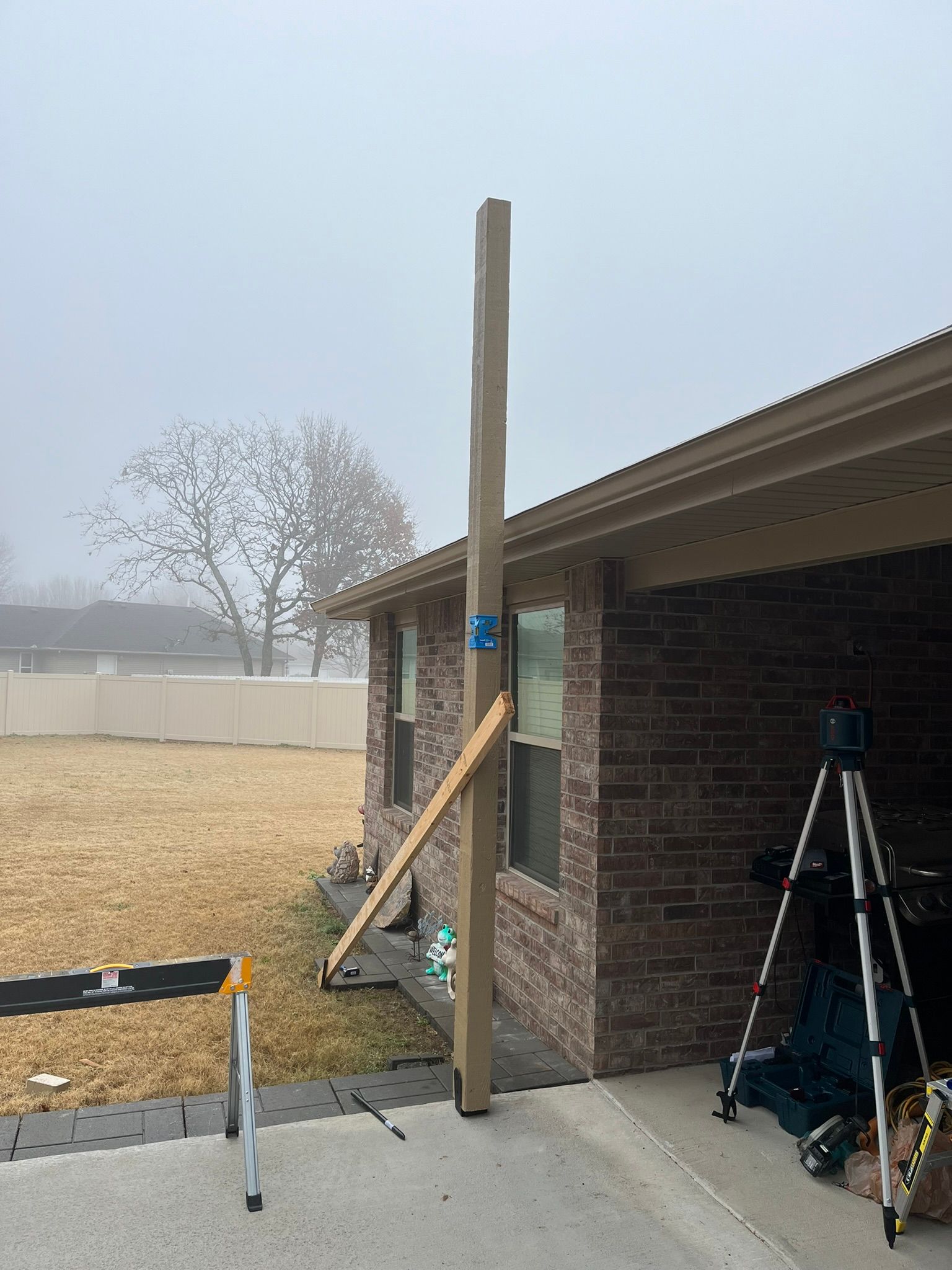 A wooden post braced against a brick building on a foggy day, with a level on a tripod in the corner.