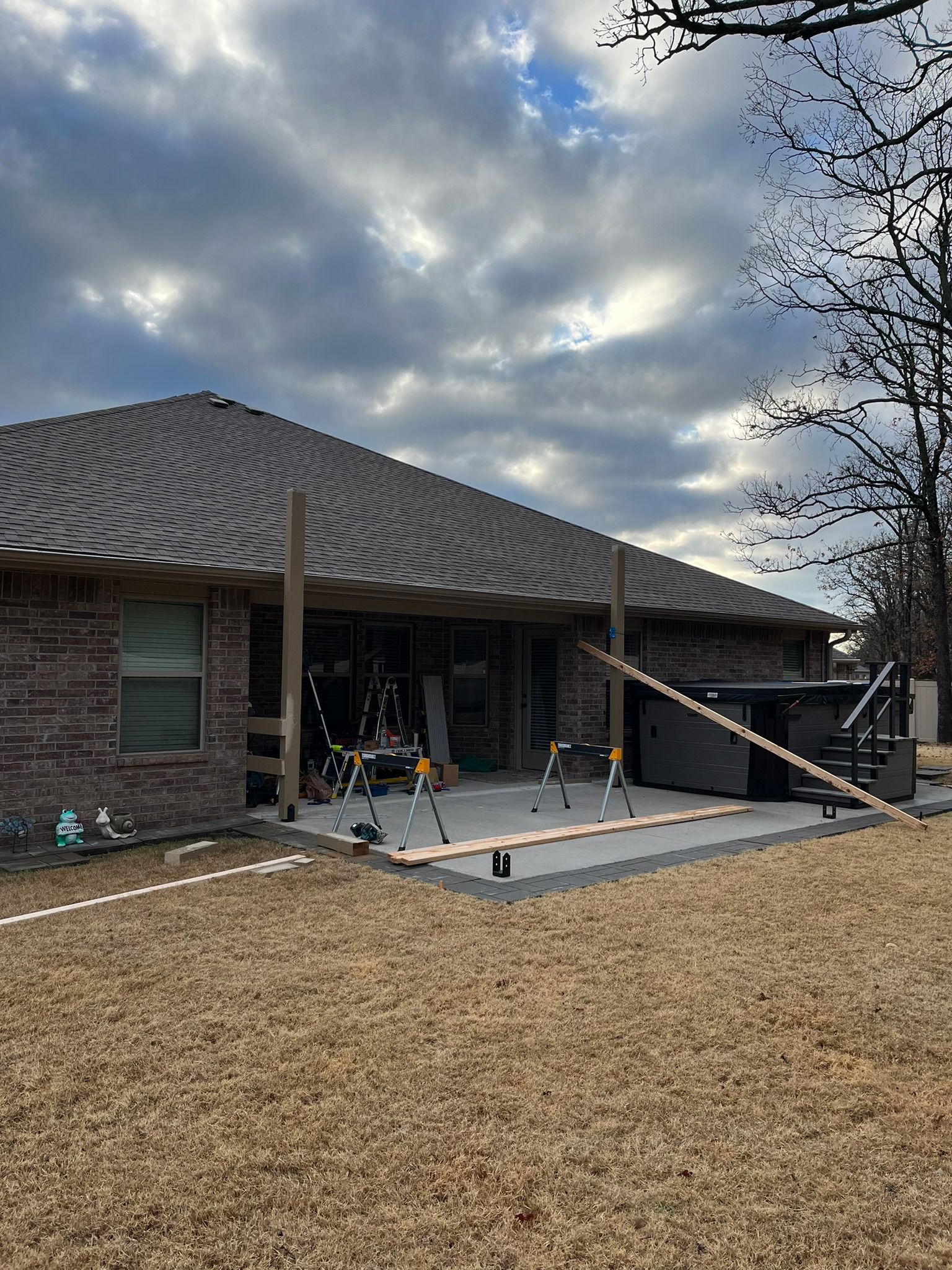 Construction of a covered patio with lumber and tools on a concrete slab near a brick house under a cloudy sky.