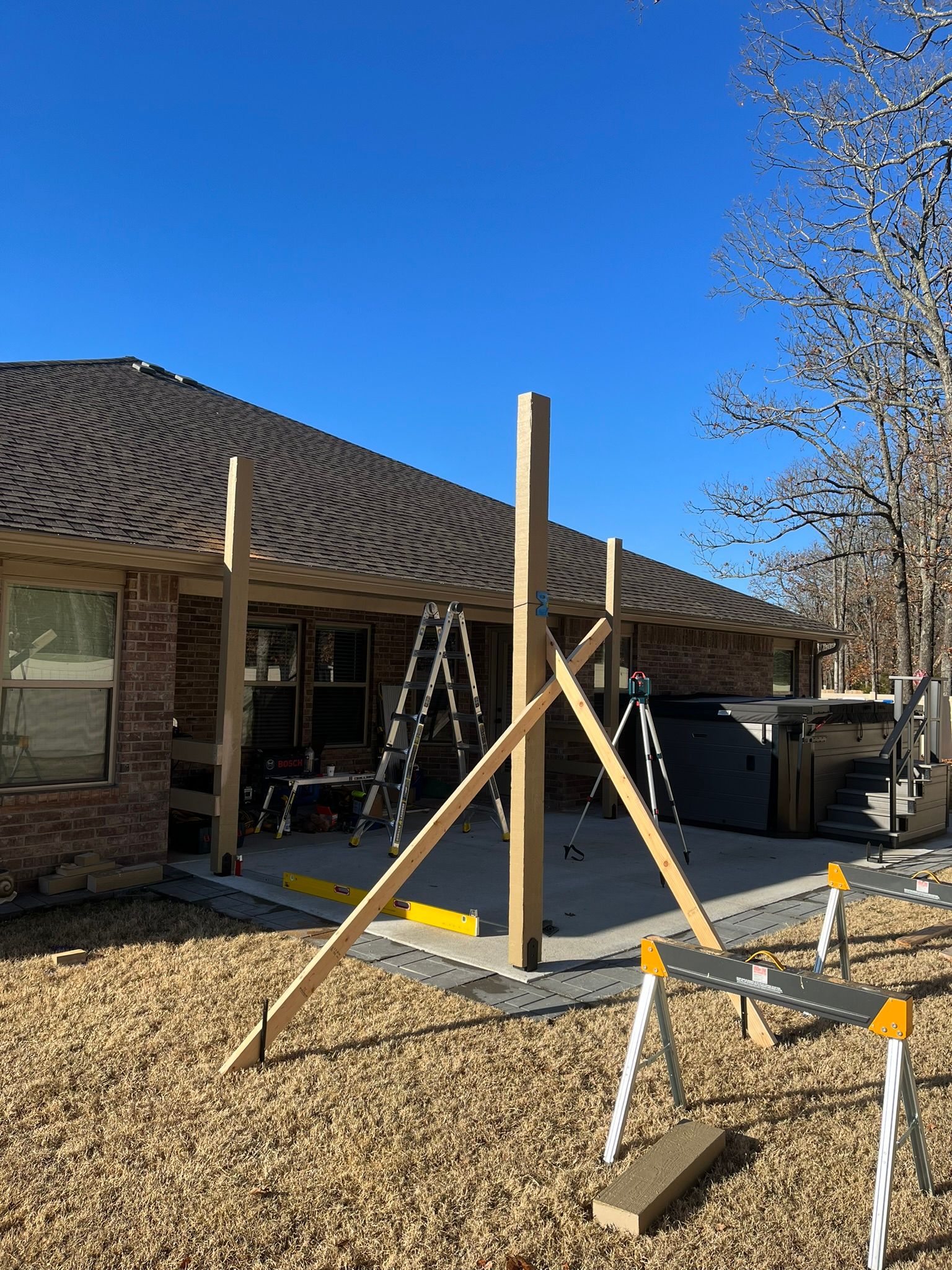 Construction of a wooden pergola on a patio near a brick house. Support beams and tools are visible. Sunny day.