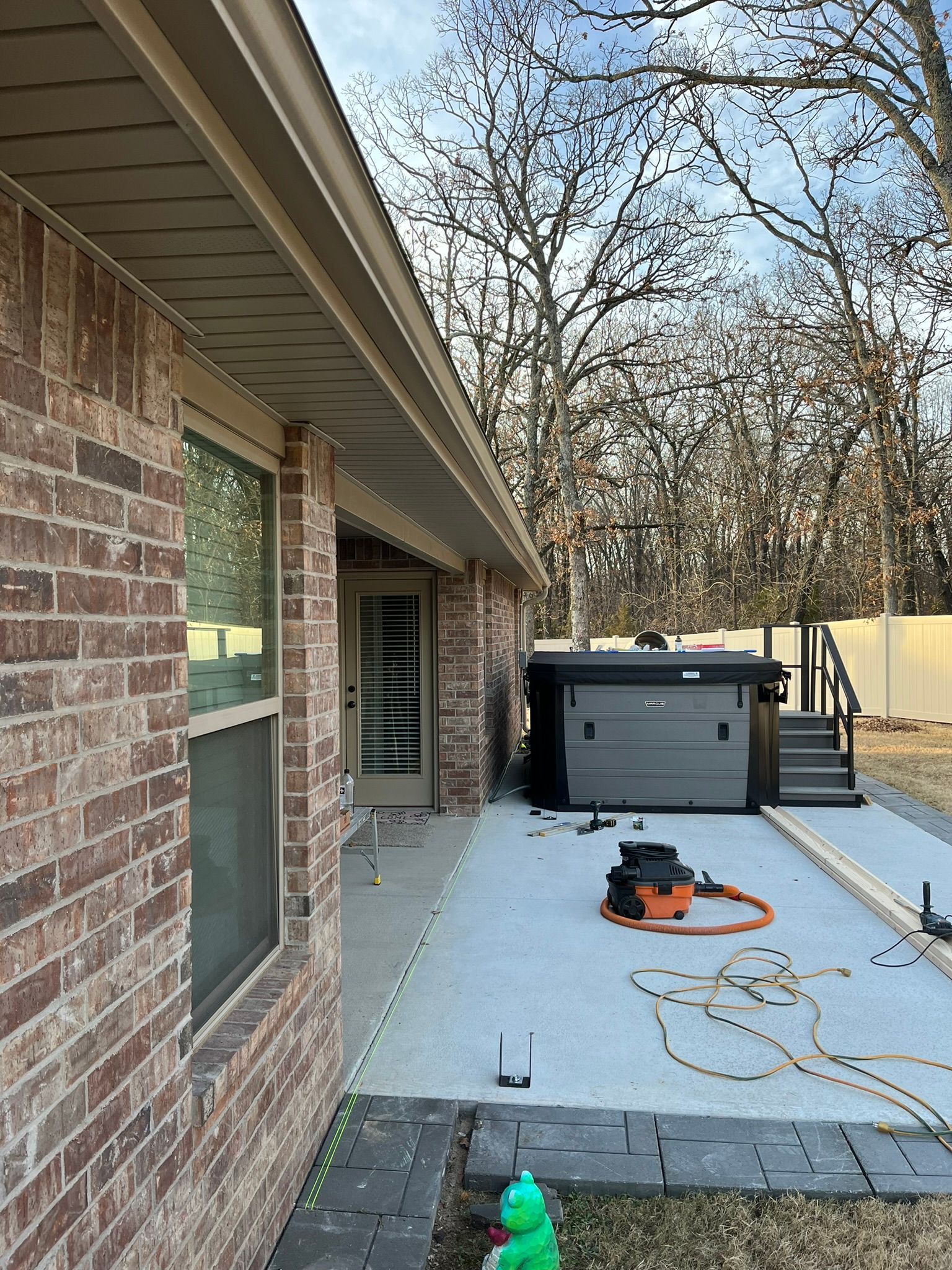 Brick house exterior with a hot tub on a concrete patio. Tools and lumber are present. Trees and sky in the background.