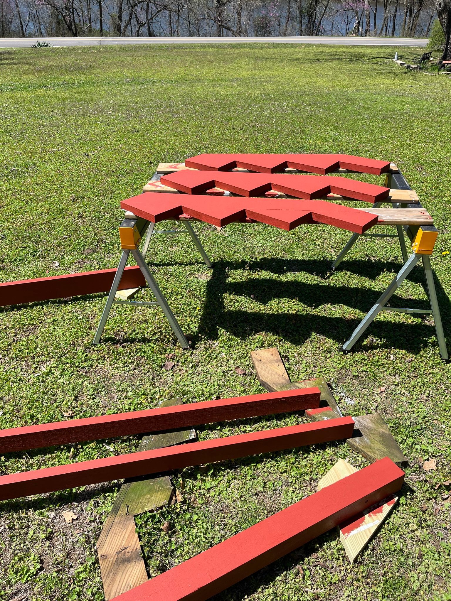 Red painted wood pieces on a sawhorse in a grassy outdoor setting, near water.