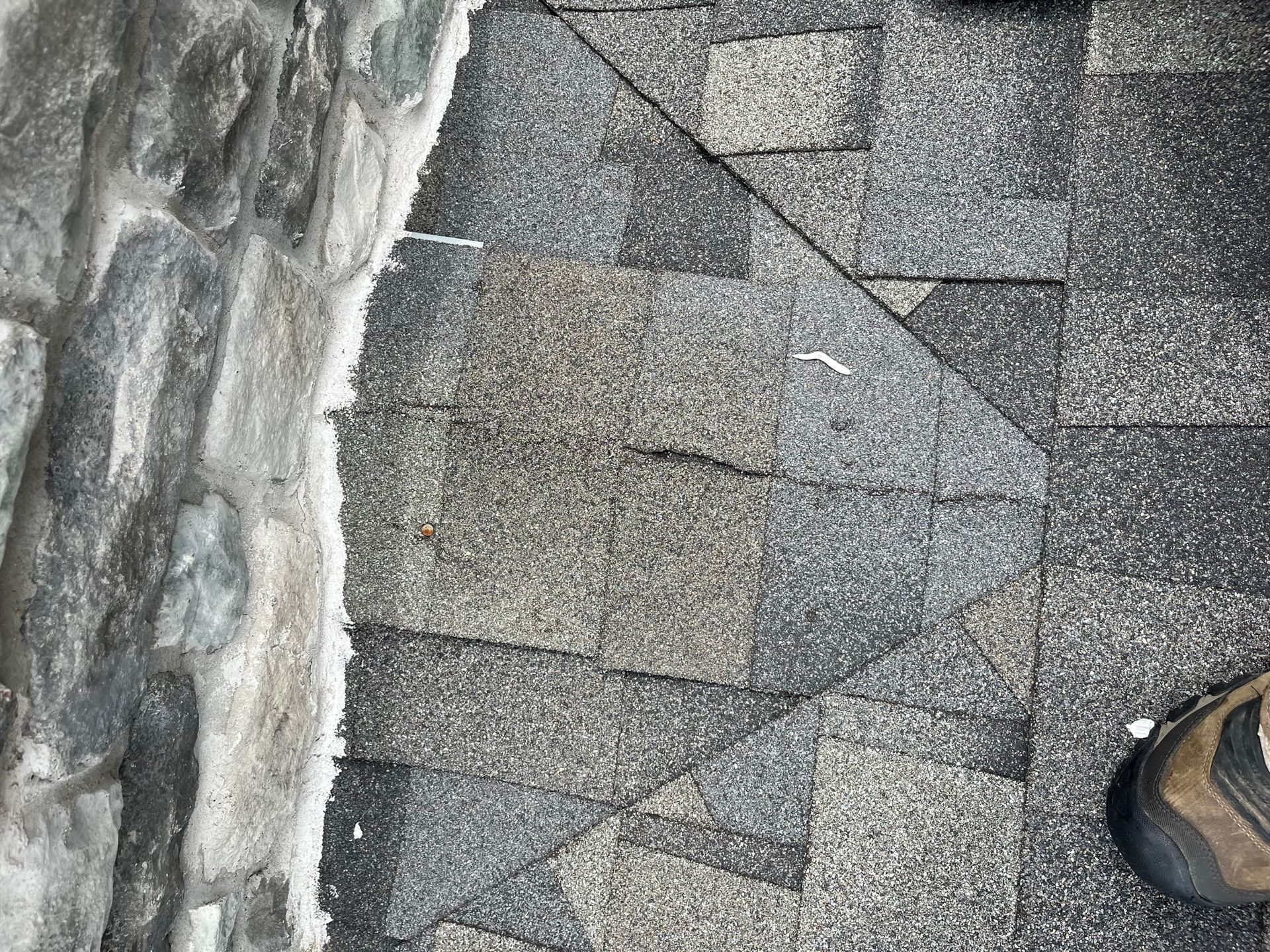 Close-up of weathered asphalt shingles next to a stone wall. A person's boot is in the frame.