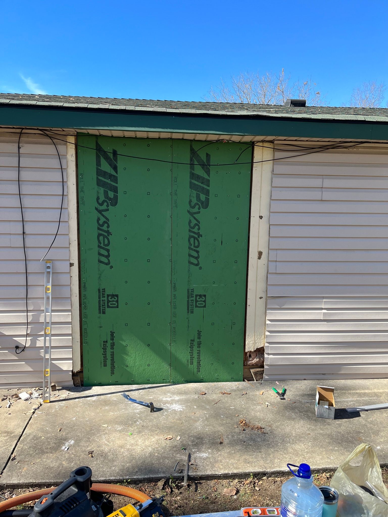 Green building sheathing covering an opening in a light-colored building wall. Concrete ground, blue sky.