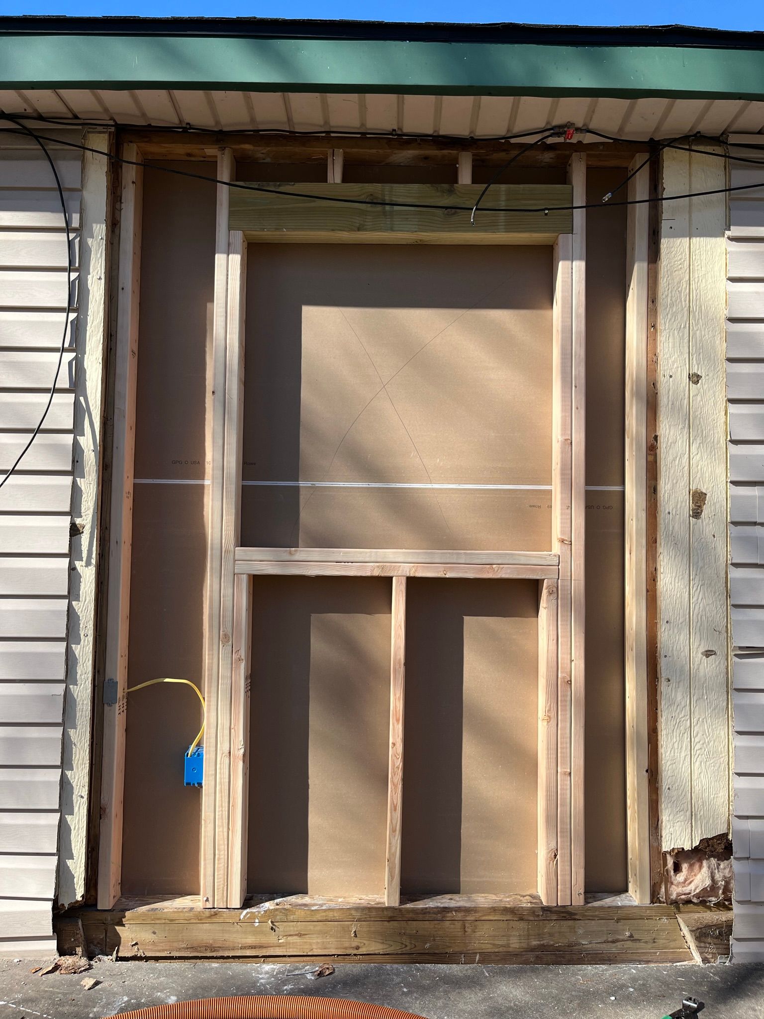 Framing of a wall opening, likely for a door. Wood framing, plywood backer, with exposed wiring and exterior siding.