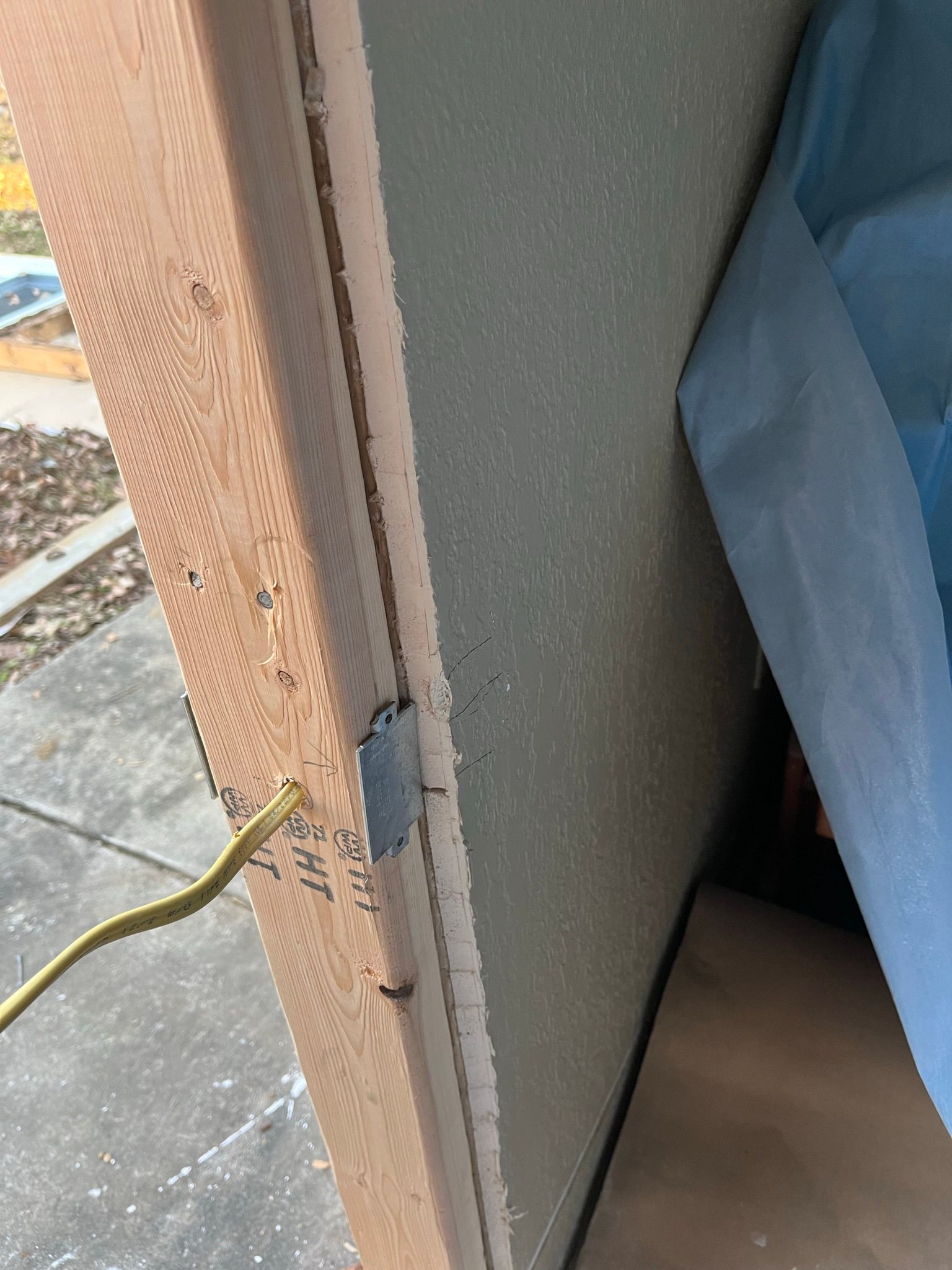 Edge of a door frame with exposed wood and wiring; exterior wall with textured surface, yellow wire.