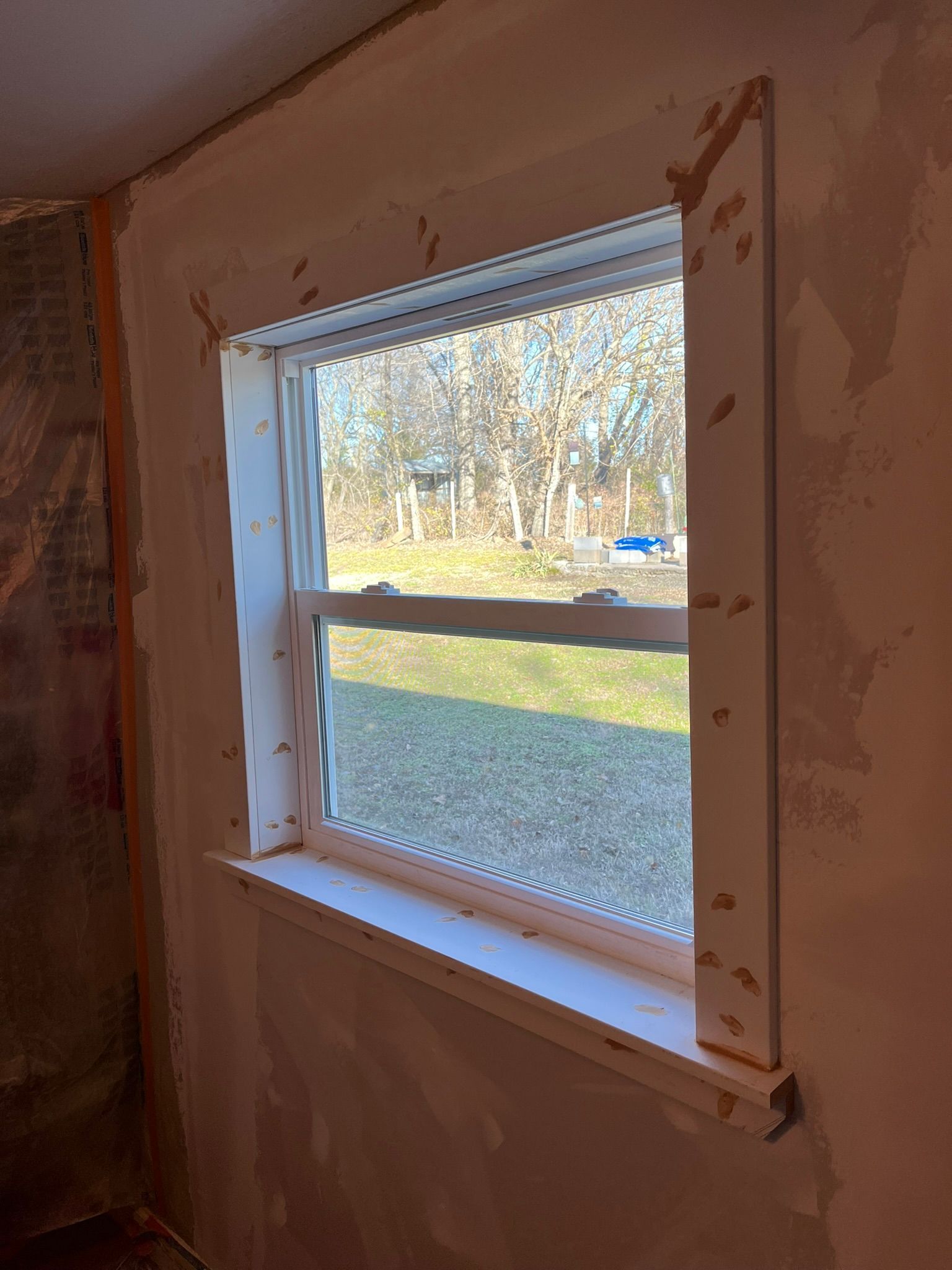 Window frame surrounded by drywall, looking out onto a grassy yard and trees.