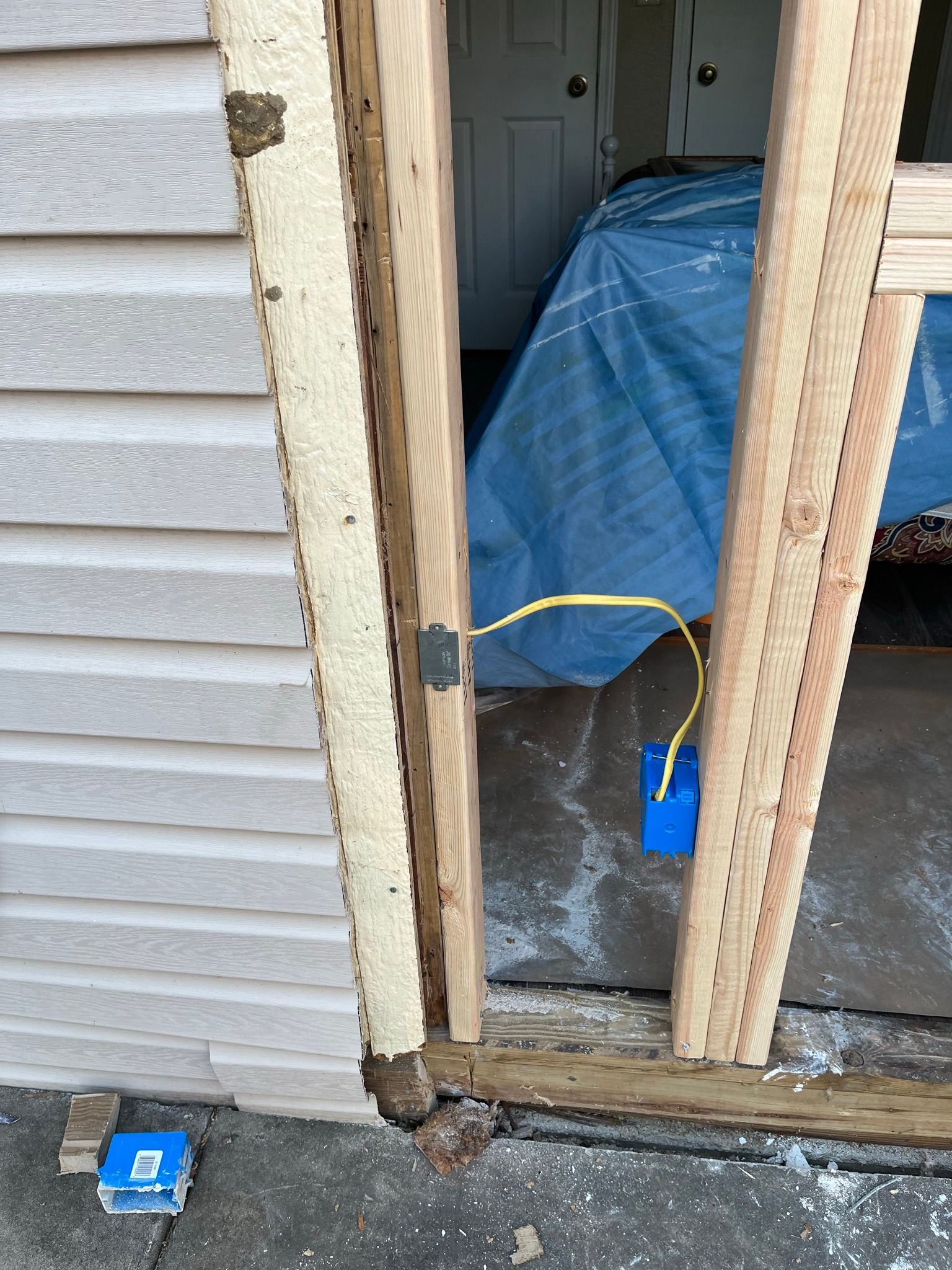 Exterior door frame under construction with exposed wiring. Beige siding, blue tarp visible.