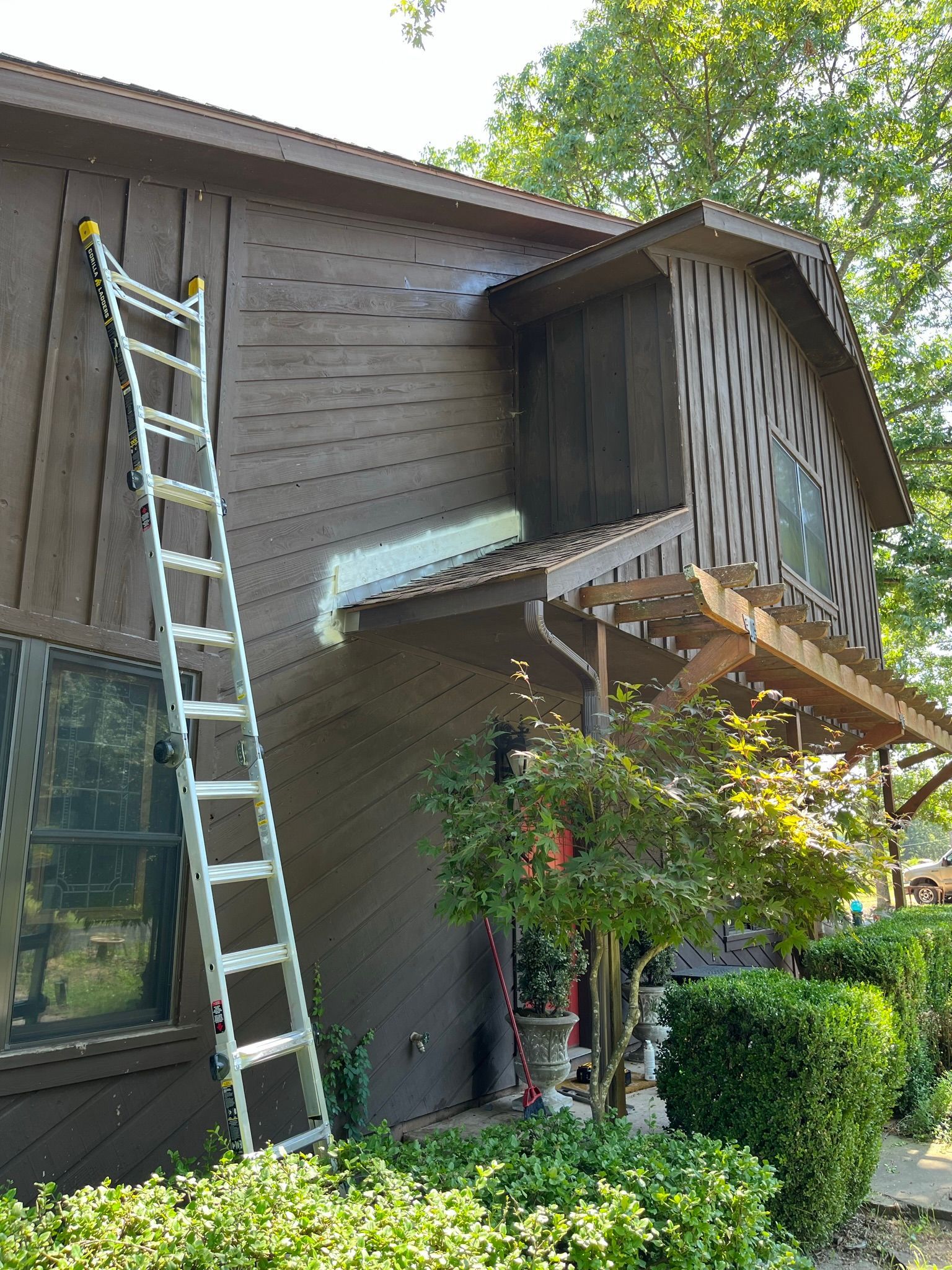 A brown house with a ladder leaning against it. Landscaping in front.