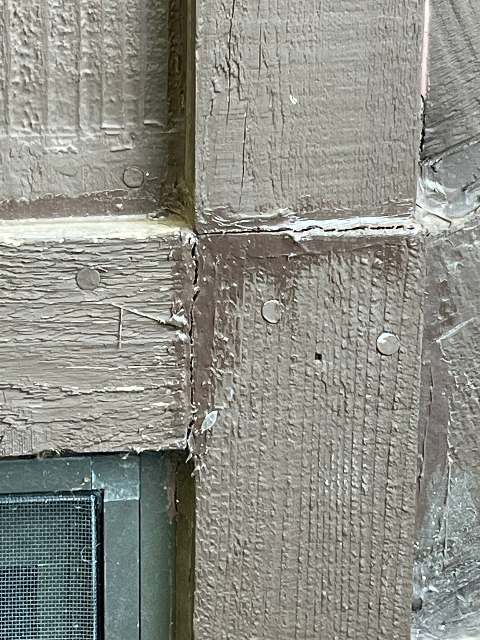 Close-up of weathered, brown painted wooden beams joined at a corner. Nails visible.