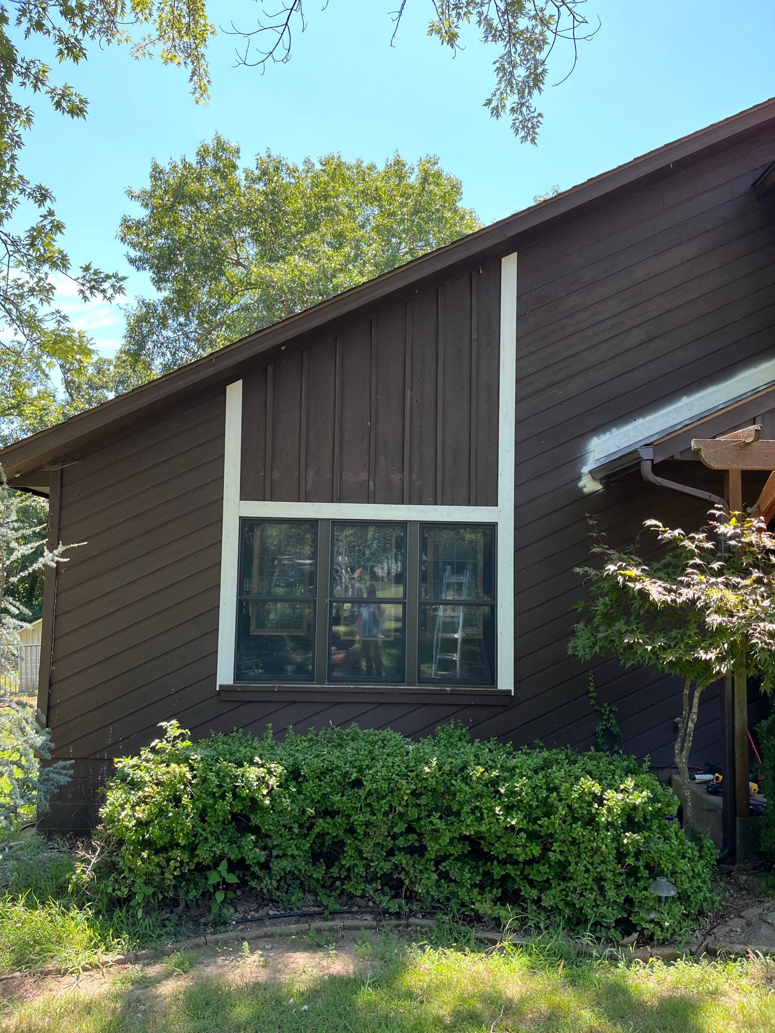Brown house with white trim around a window, green bushes in front.