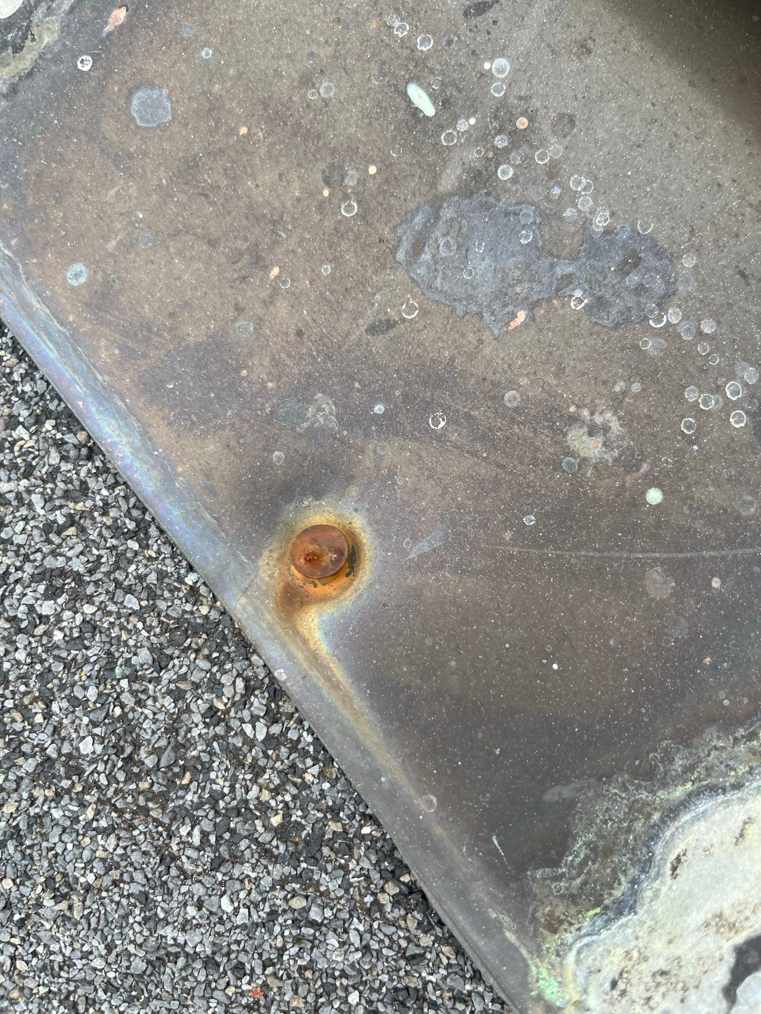 Close-up of a weathered, dark metal surface with a rusty, circular detail. Surface is next to gray gravel.