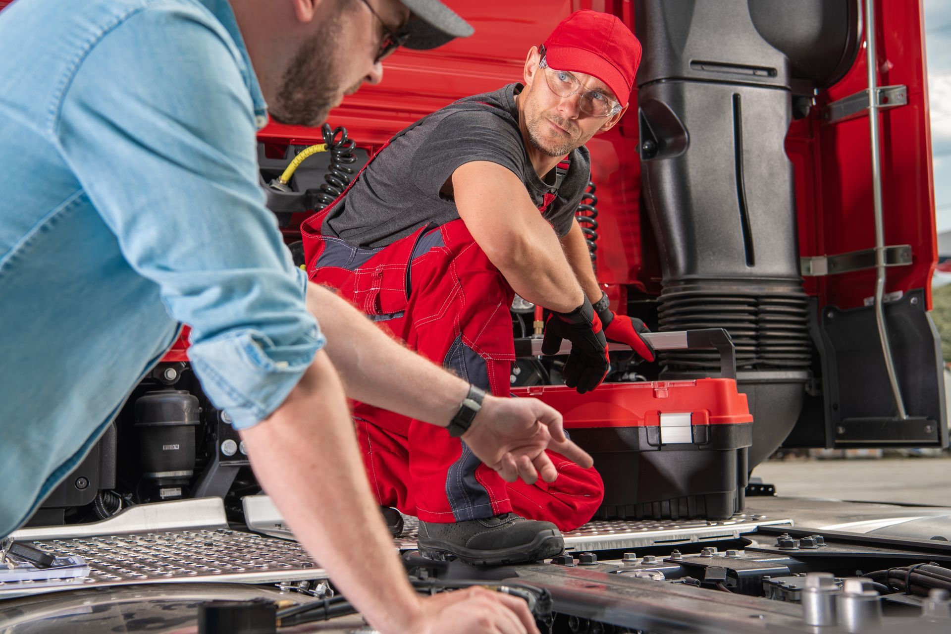 Two mechanics working on a vehicle engine in a garage, one holding a wrench in a red cap.