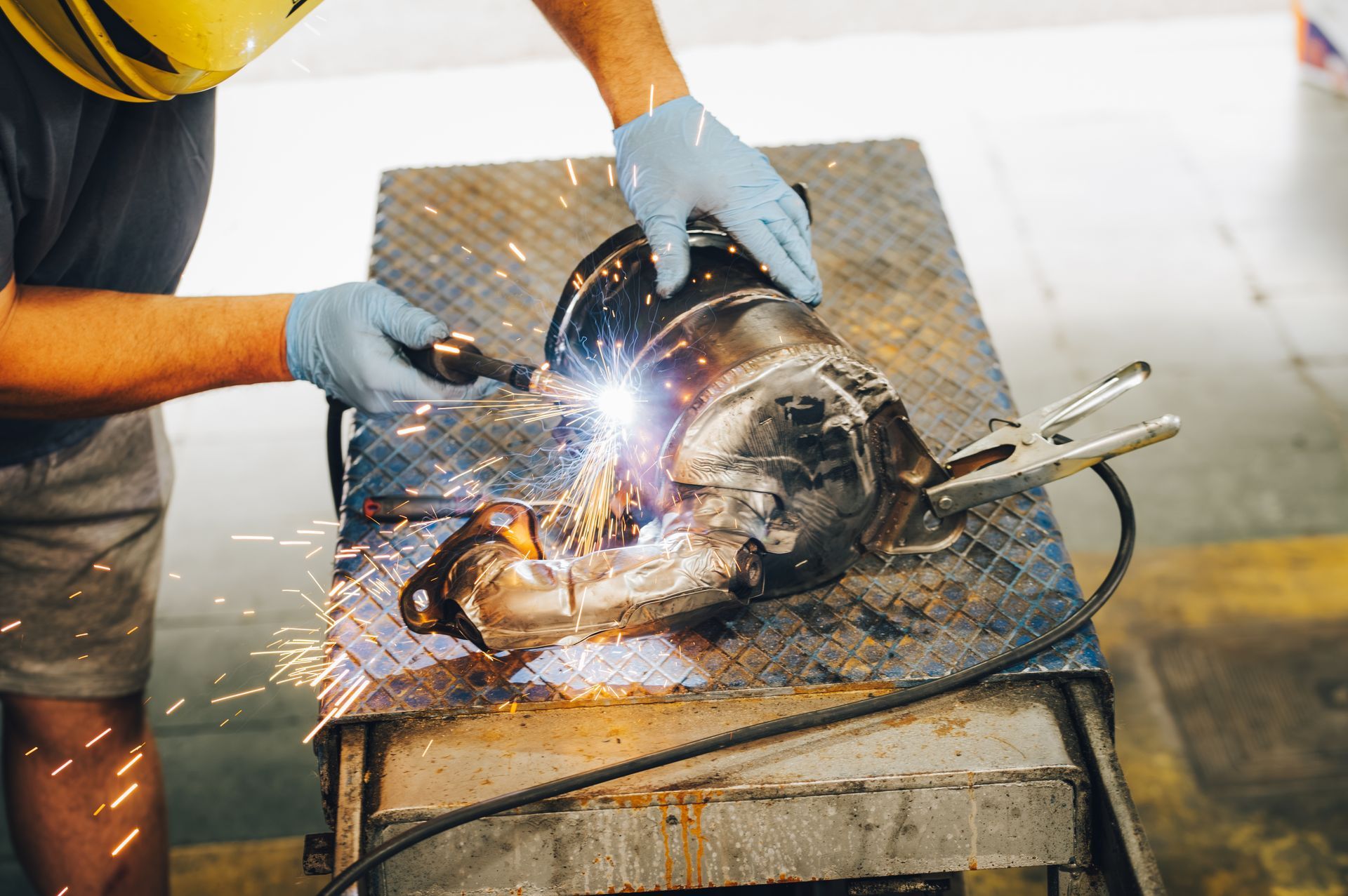 Welder sparks on a metal part in a workshop, wearing blue gloves and a yellow hard hat