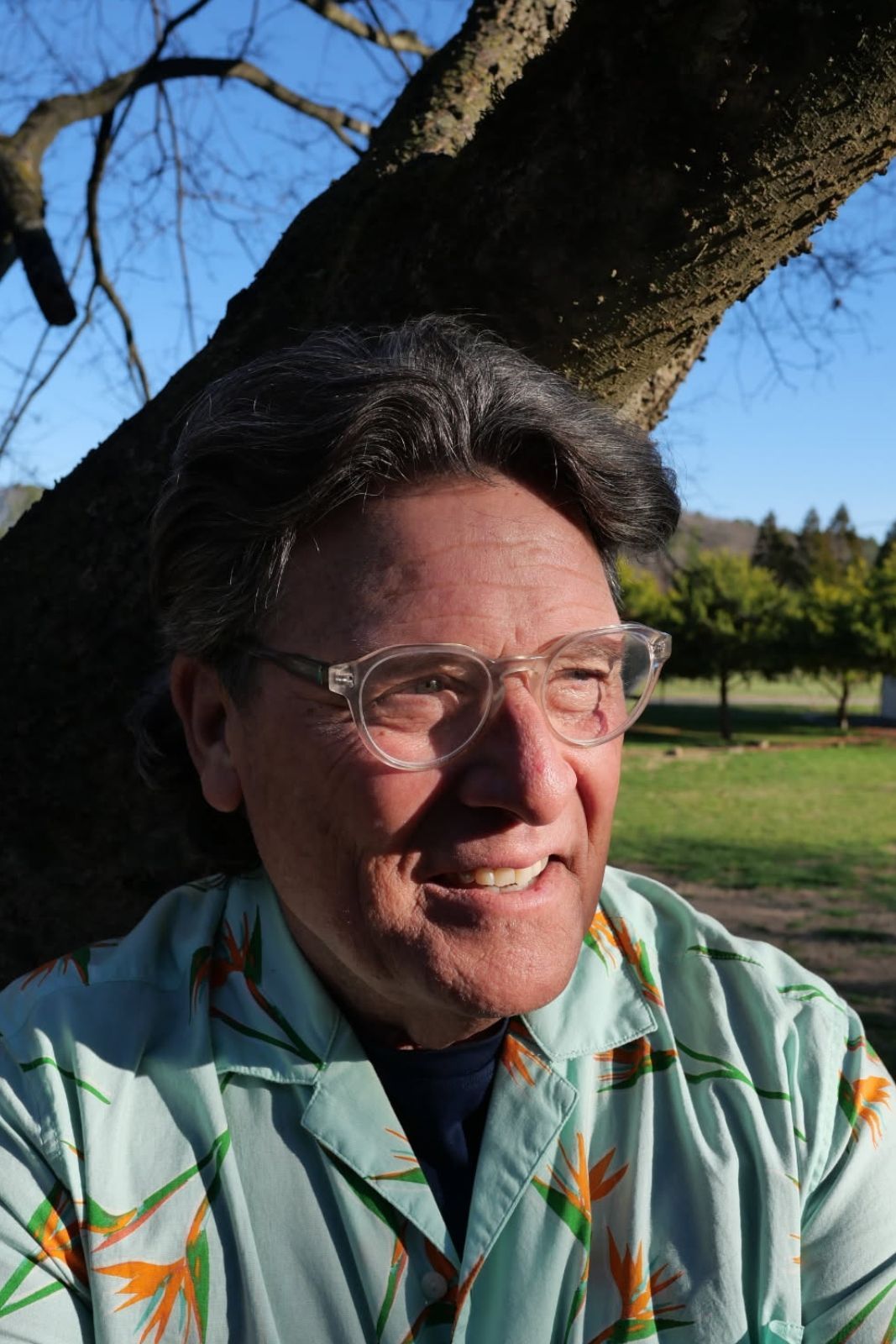 Man with clear glasses smiles outdoors, leaning against a tree. He wears a light blue floral shirt.