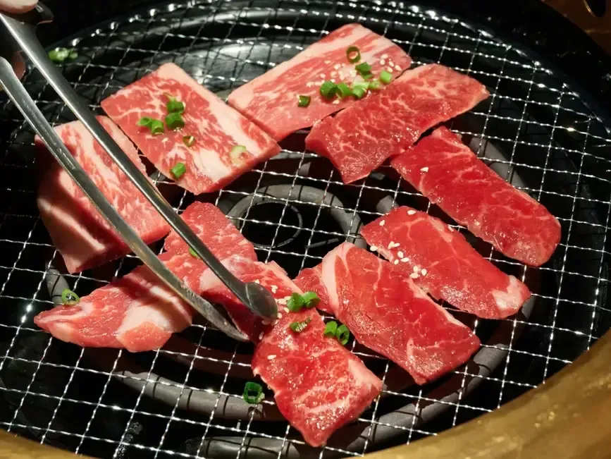 Raw marinated beef slices grilling on a wire rack, with tongs turning the meat over charcoal.