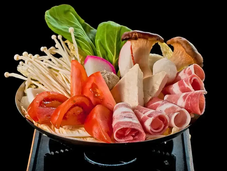 Hot pot with assorted meats, mushrooms, greens, and sliced tomatoes in a black bowl