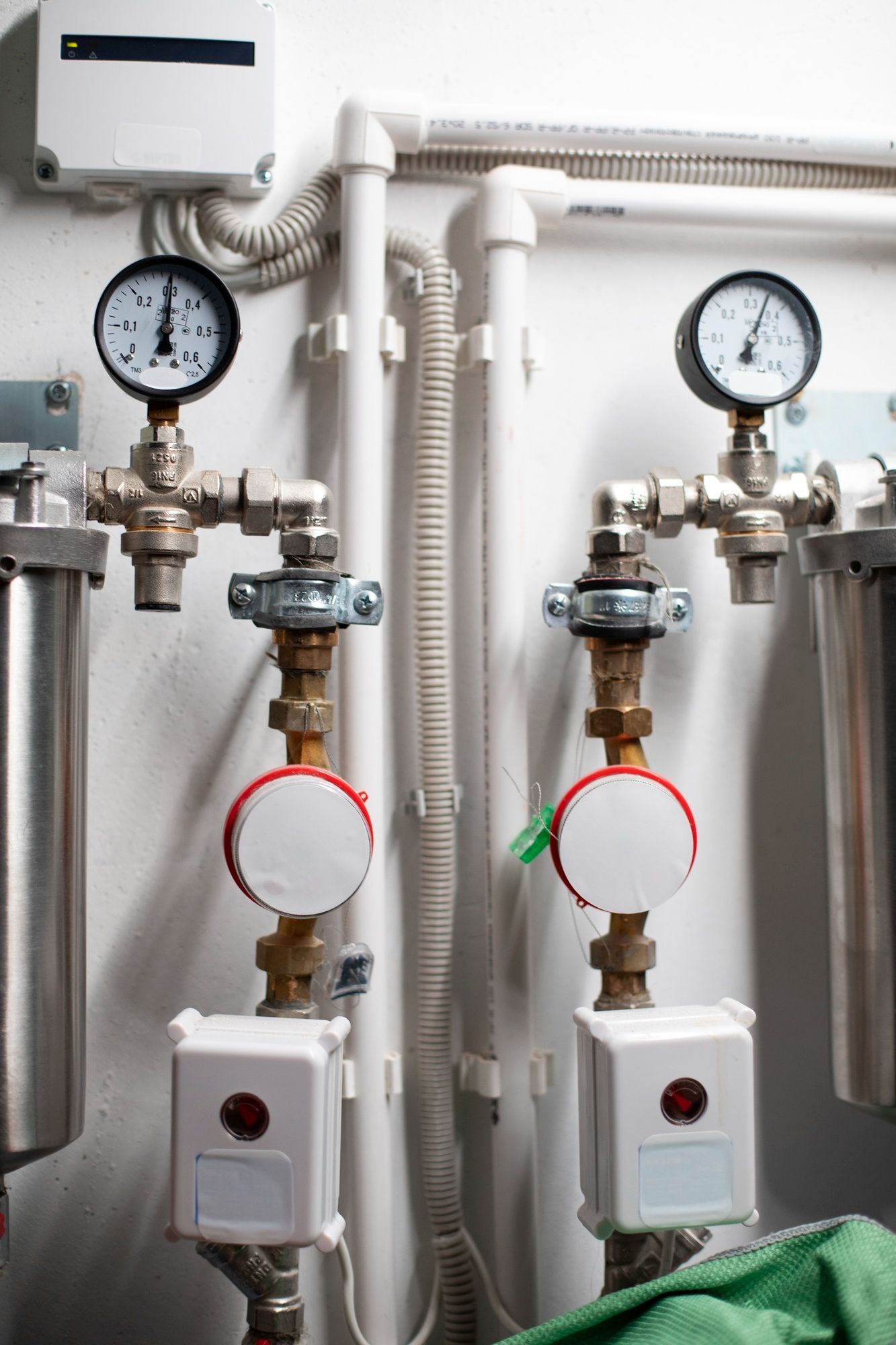 Two industrial water meters with pressure gauges, circular dial indicators, and small sensors attached to white wall pipes.