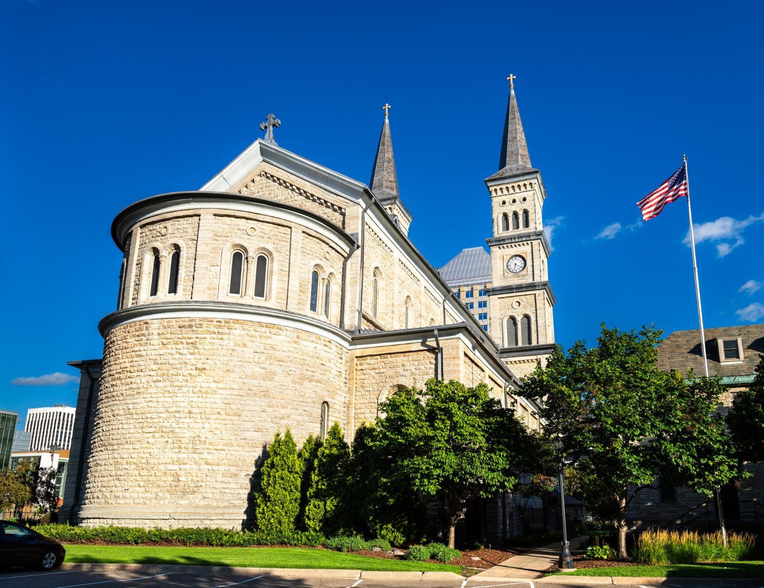 A historic stone church with two spires and a rounded apse under a clear blue sky, with an American flag flying nearby.