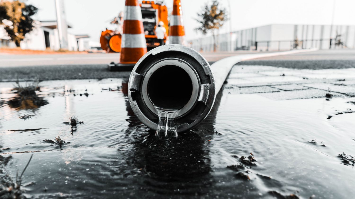 A metal hose nozzle rests on a wet, paved surface with water dripping from it, near orange traffic cones.