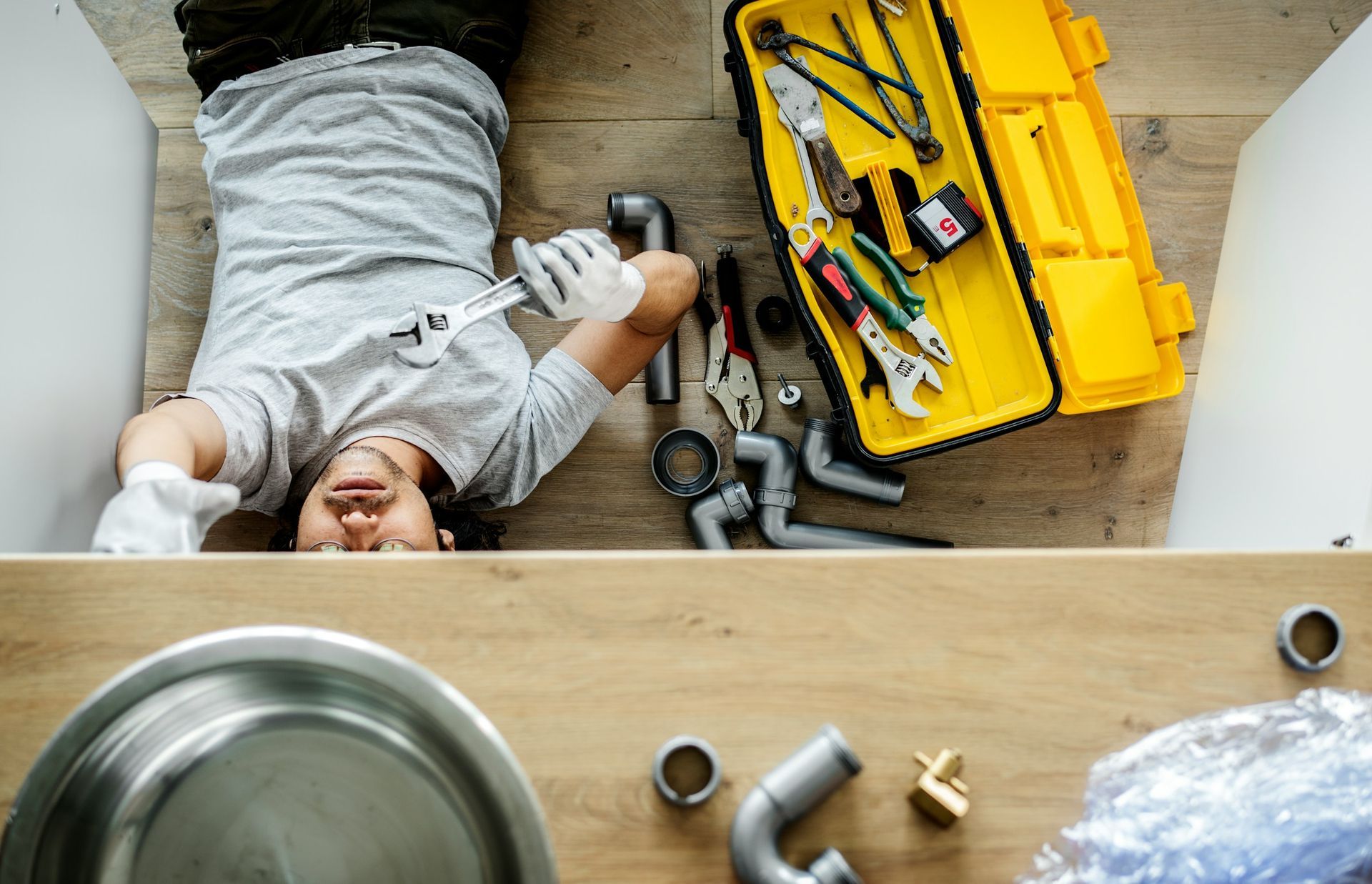 A person lying on the floor under a kitchen sink, wearing gloves and using a wrench to repair plumbing components.