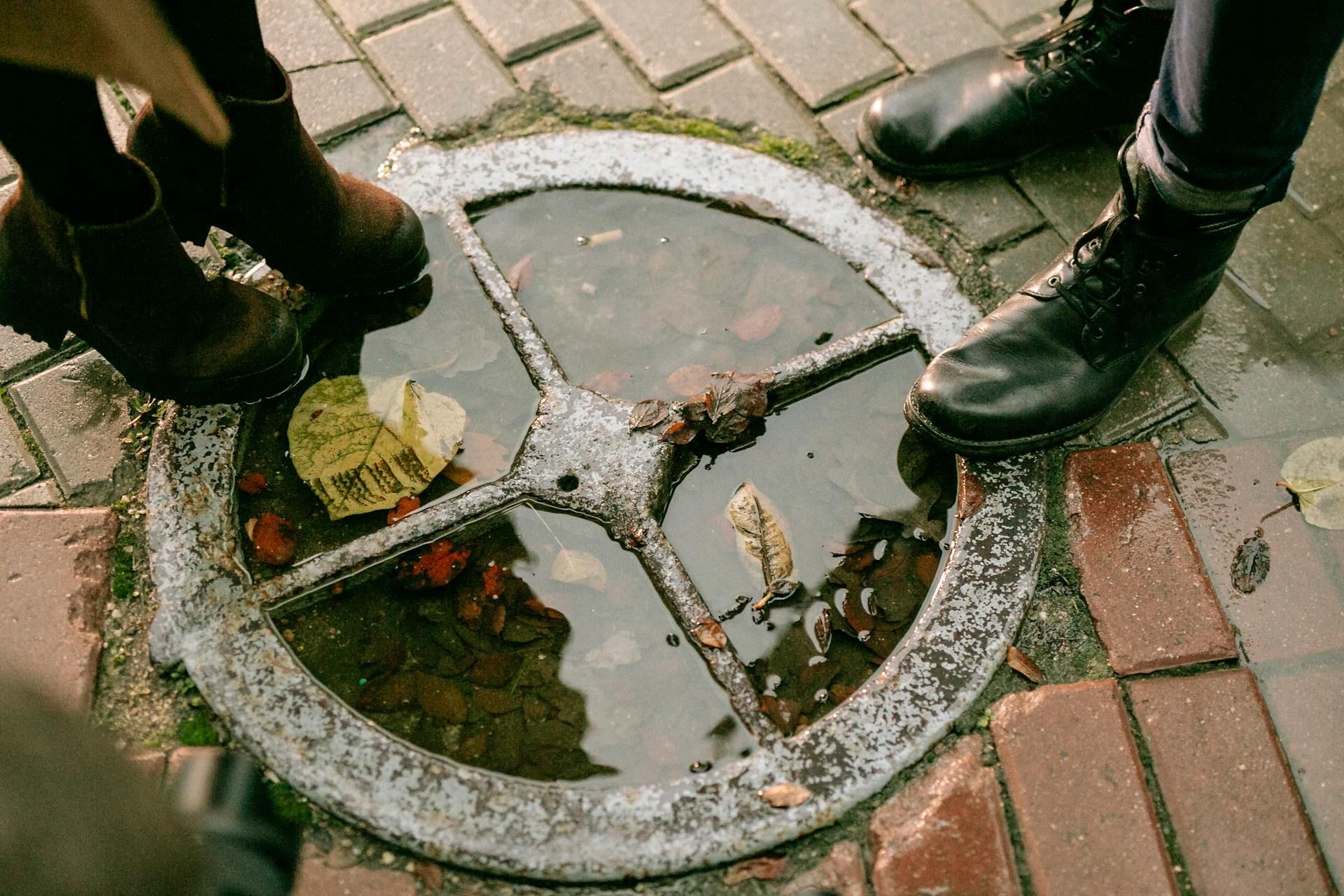 Feet in dark boots stand on a paved surface next to a circular metal grate filled with rainwater and fallen leaves.